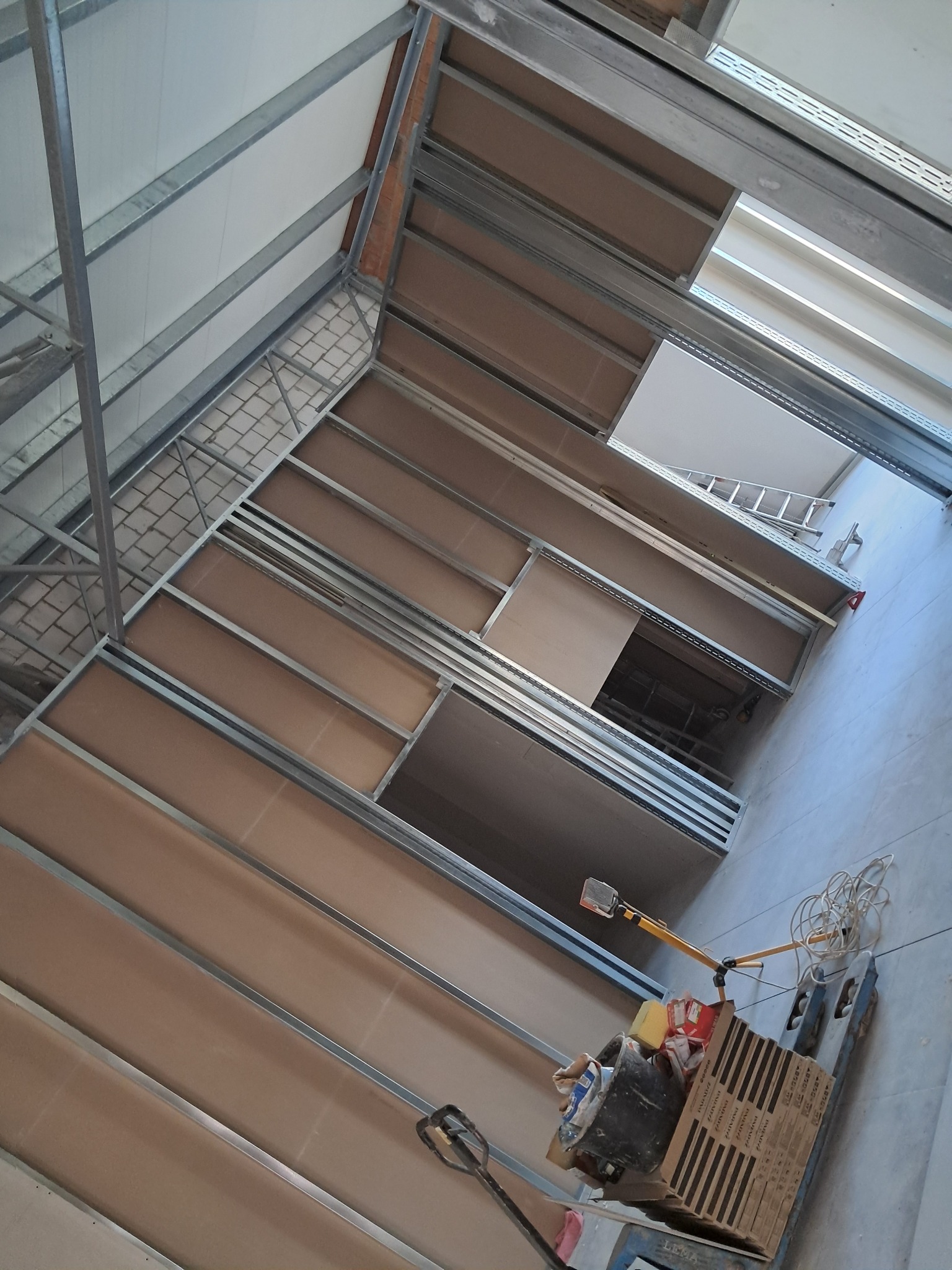 Konstrukcja metalowa pod zabudowę poddasza z płytami karton-gips, widoczny wózek transportowy z narzędziami i materiałami budowlanymi, w tle drabina aluminiowa.