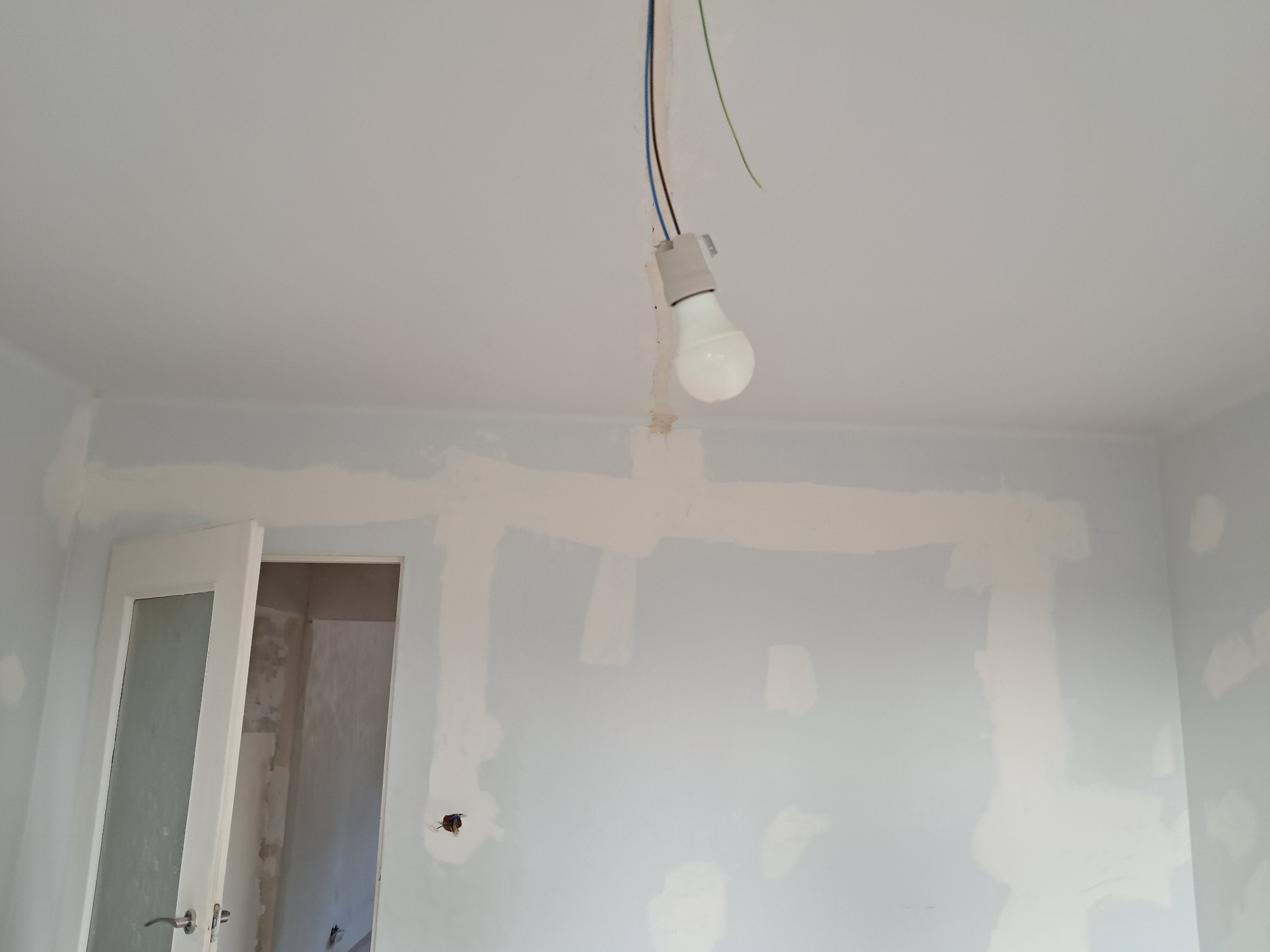 Pokój w trakcie prac: ściany z nałożoną gładzią miejscami, wisząca żarówka na kablu, otwarte drzwi do kolejnego pomieszczenia.
