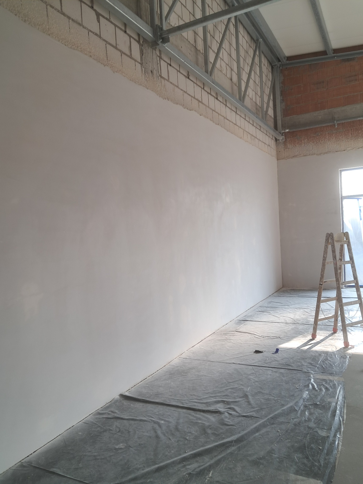 Świeżo pomalowana, jasna ściana w surowym wnętrzu z widoczną konstrukcją stropu i ceglanymi fragmentami. Podłoga zabezpieczona folią malarską, w tle drabina.