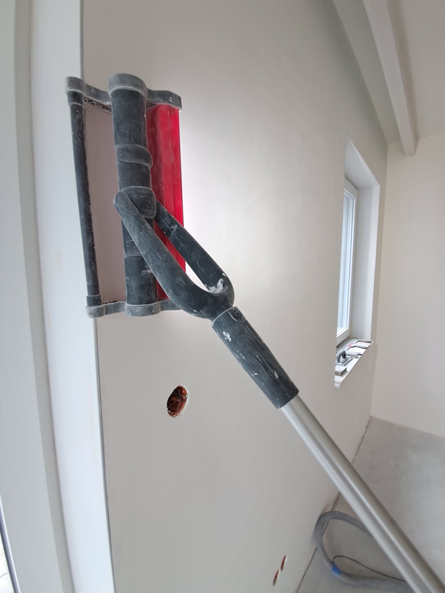 Szlifowanie ściany szlifierką z długim uchwytem, widoczny otwór na gniazdko elektryczne i okno z narzędziami na parapecie.