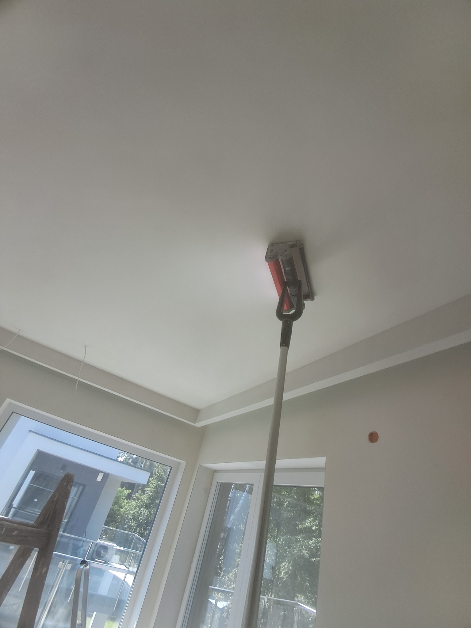 Szlifowanie sufitu za pomocą szlifierki na wysięgniku w pomieszczeniu z oknami i listwami dekoracyjnymi.