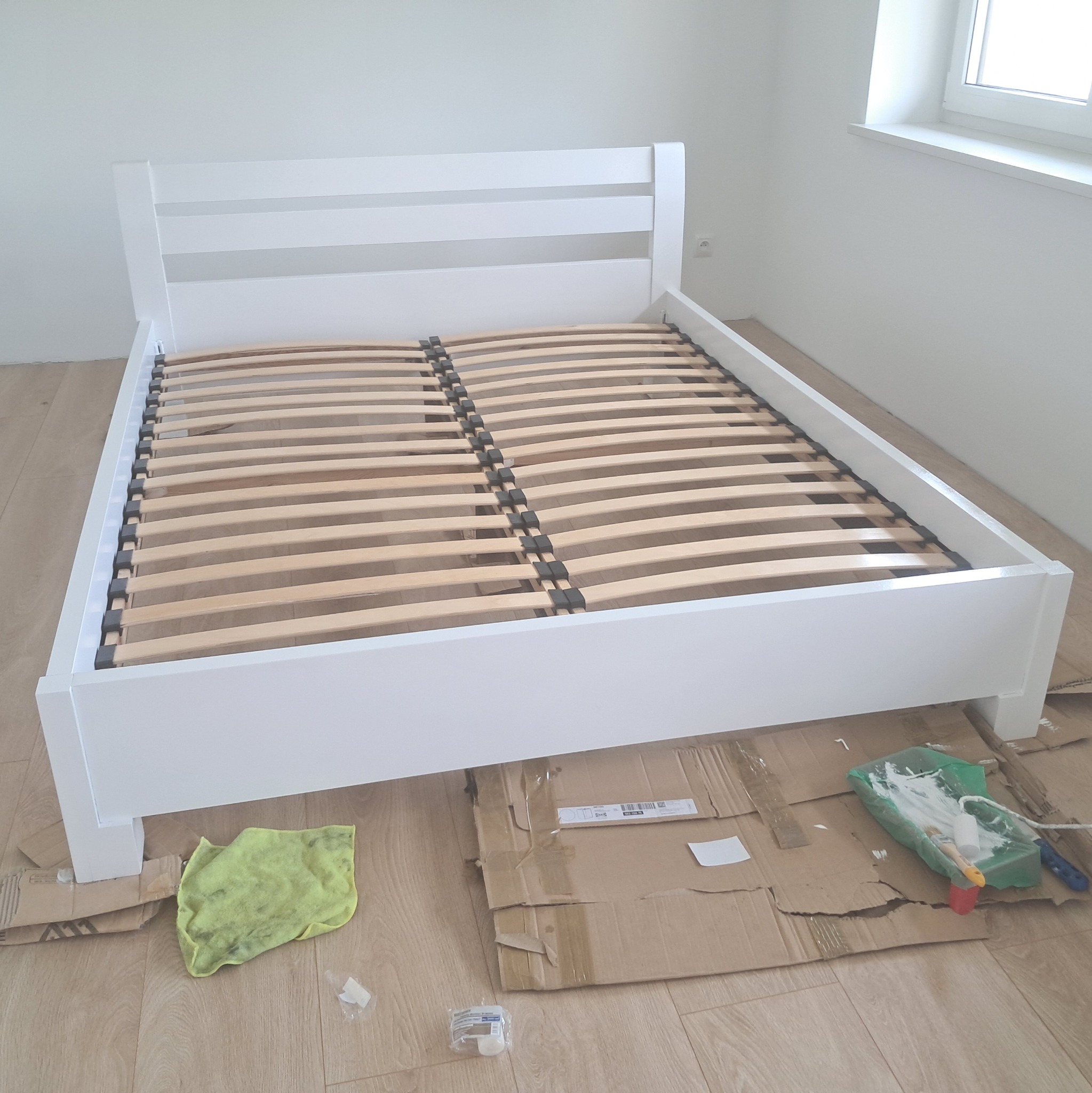 Białe łóżko z drewnianym stelażem w trakcie malowania, zabezpieczone kartonem, z widocznym wałkiem i pędzlem malarskim w kuwecie z farbą.