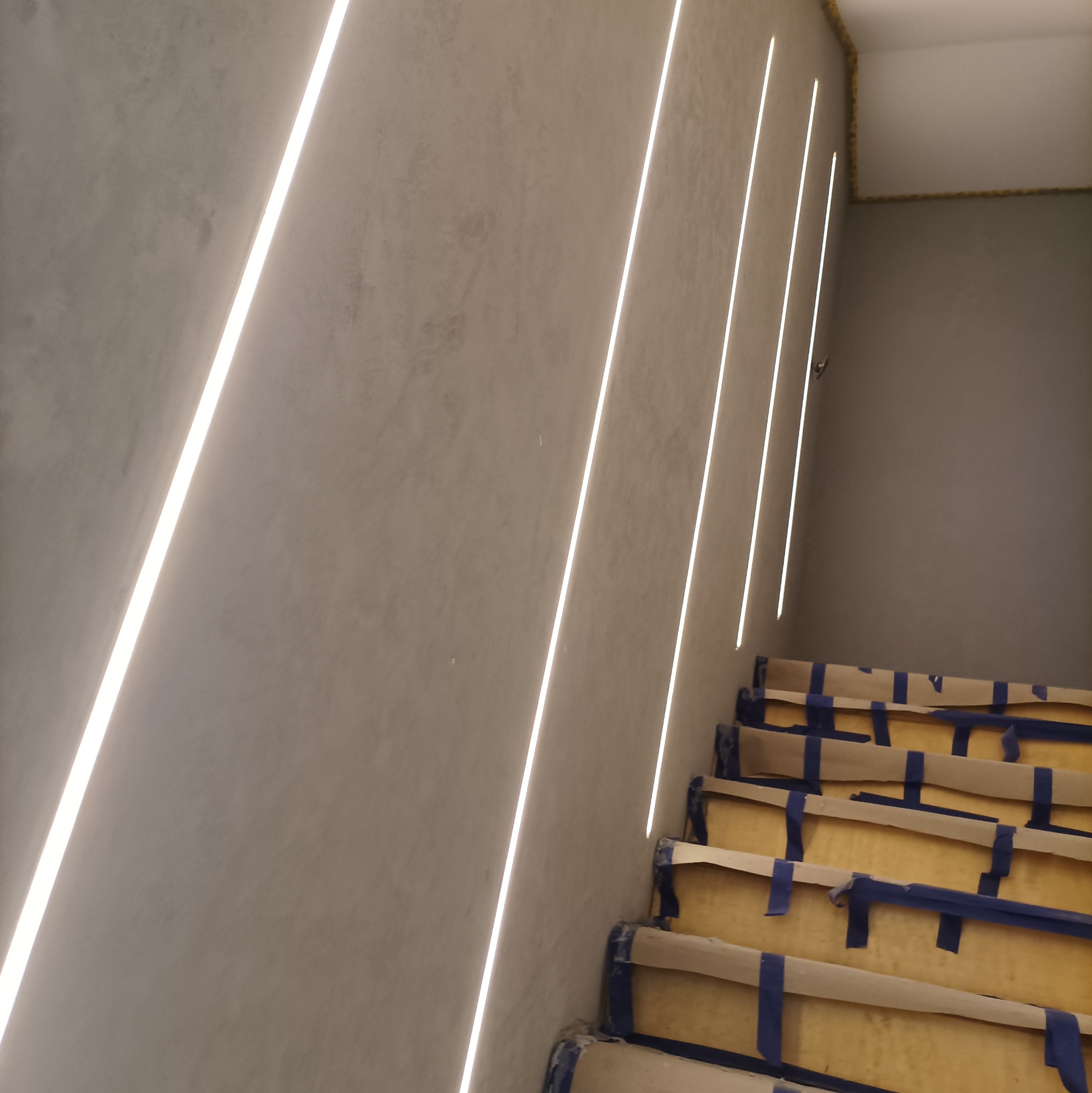 Ściana ze strukturą betonu architektonicznego z wpuszczonymi pionowymi liniami świetlnymi LED, schody zabezpieczone kartonem i taśmą malarską podczas prac.