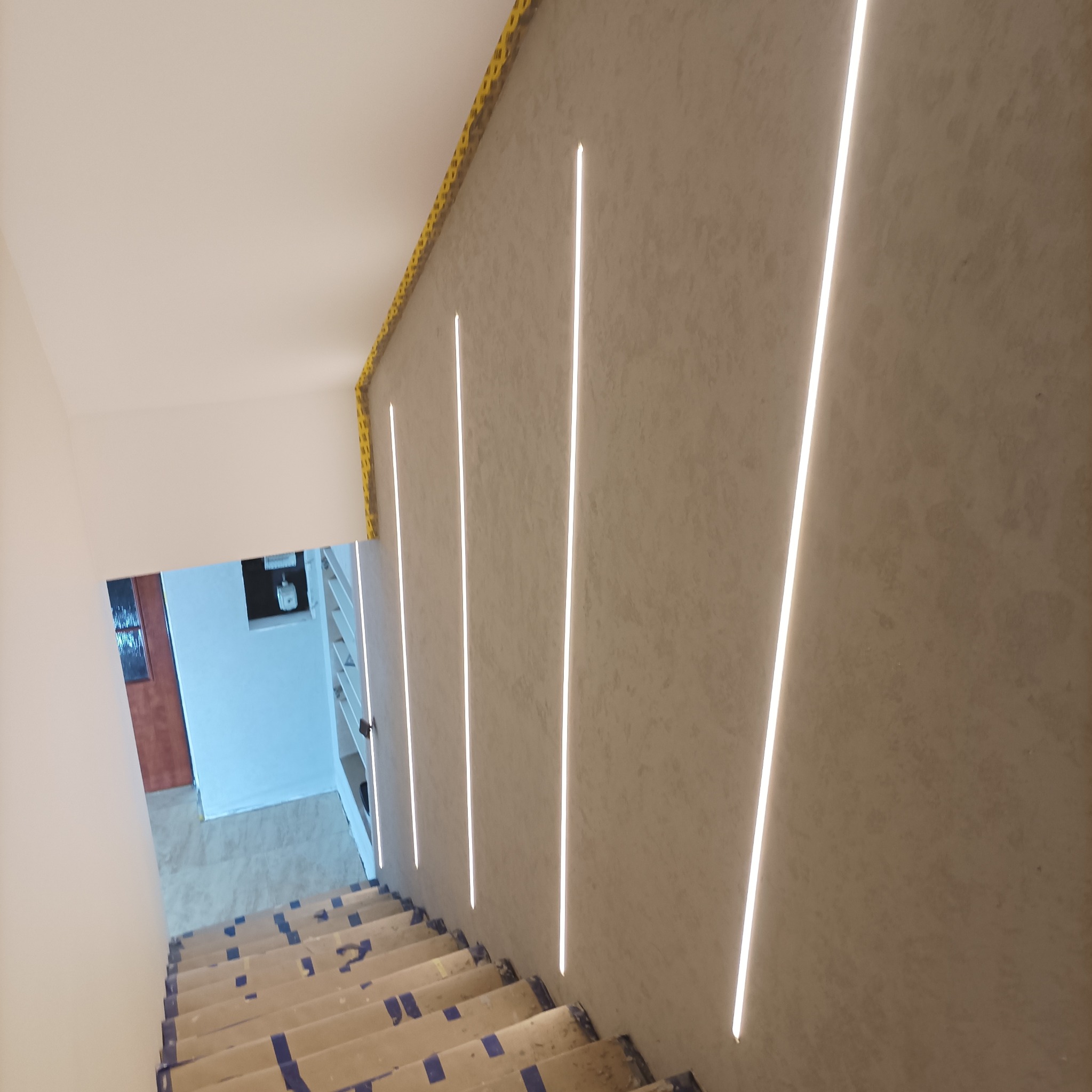 Ściana wykończona gładzią z wpuszczonymi pionowymi listwami LED, schody zabezpieczone folią malarską podczas prac wykończeniowych.