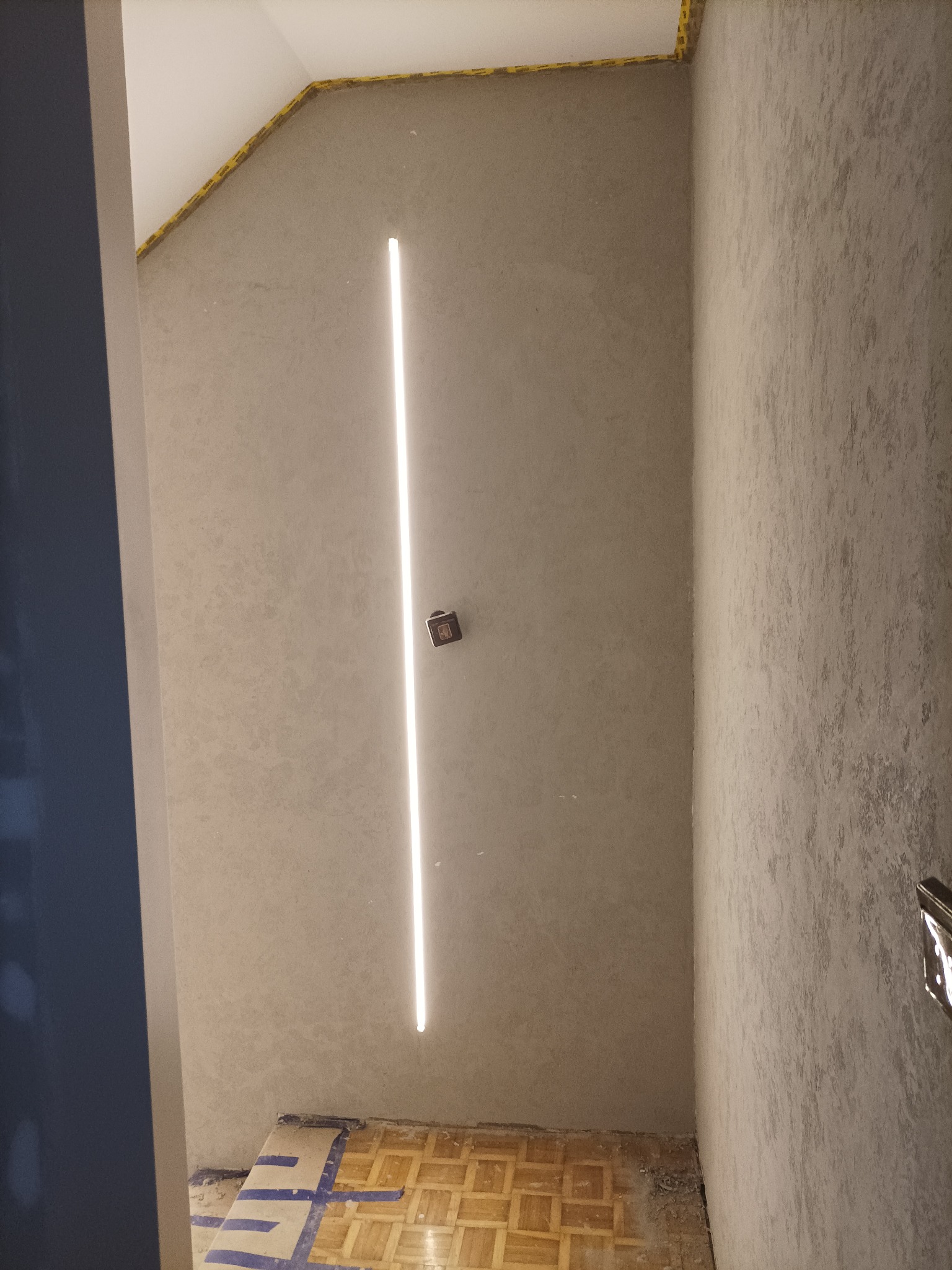 Ściana po nałożeniu gładzi z zamontowanym oświetleniem LED, widoczne zabezpieczenia taśmą malarską przy suficie i podłodze, fragment parkietu.