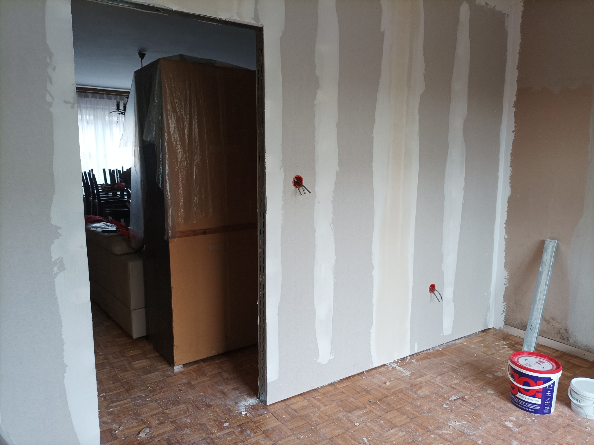 Ściana w trakcie prac przygotowawczych, z widocznymi pionowymi paskami świeżo nałożonej masy szpachlowej, wystającymi przewodami elektrycznymi, zabezpieczonym meblem w tle oraz wiadrami z farbą...