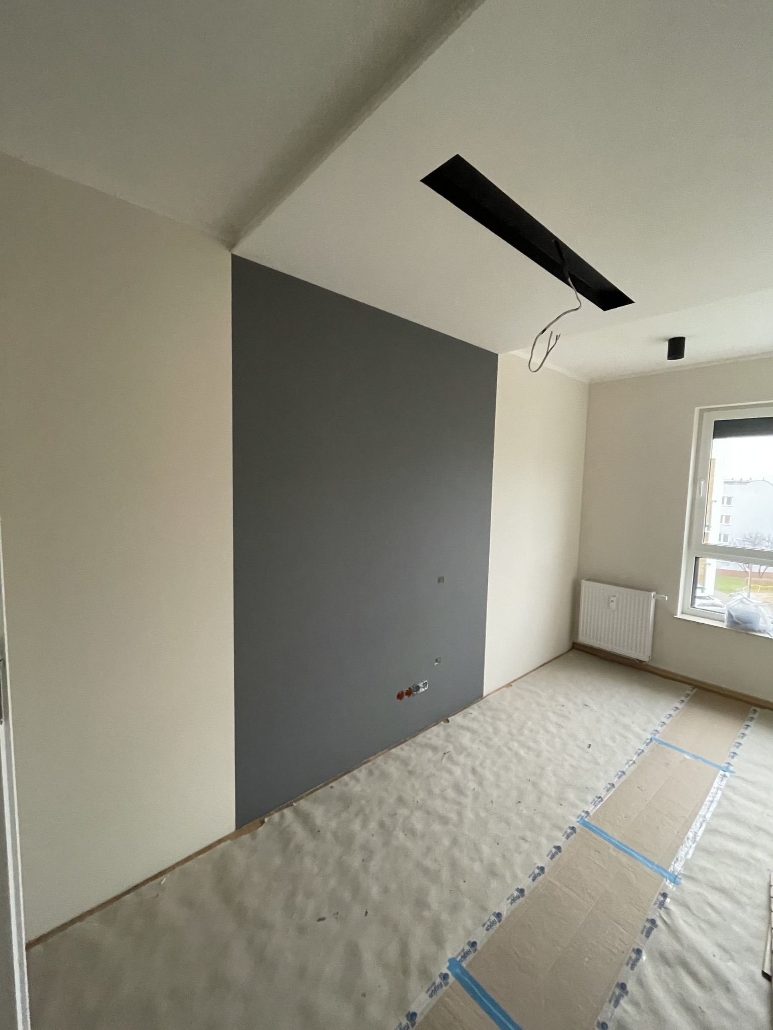 Pokój po częściowym malowaniu: jedna ściana pomalowana na szaro, reszta na beżowo, podłoga zabezpieczona folią malarską, widoczne gniazdka elektryczne i puszka instalacyjna w suficie.