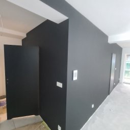 Świeżo pomalowane na czarno wnętrze z widocznymi gniazdkami elektrycznymi i ościeżnicą drzwiową. Podłoga częściowo zabezpieczona folią malarską.