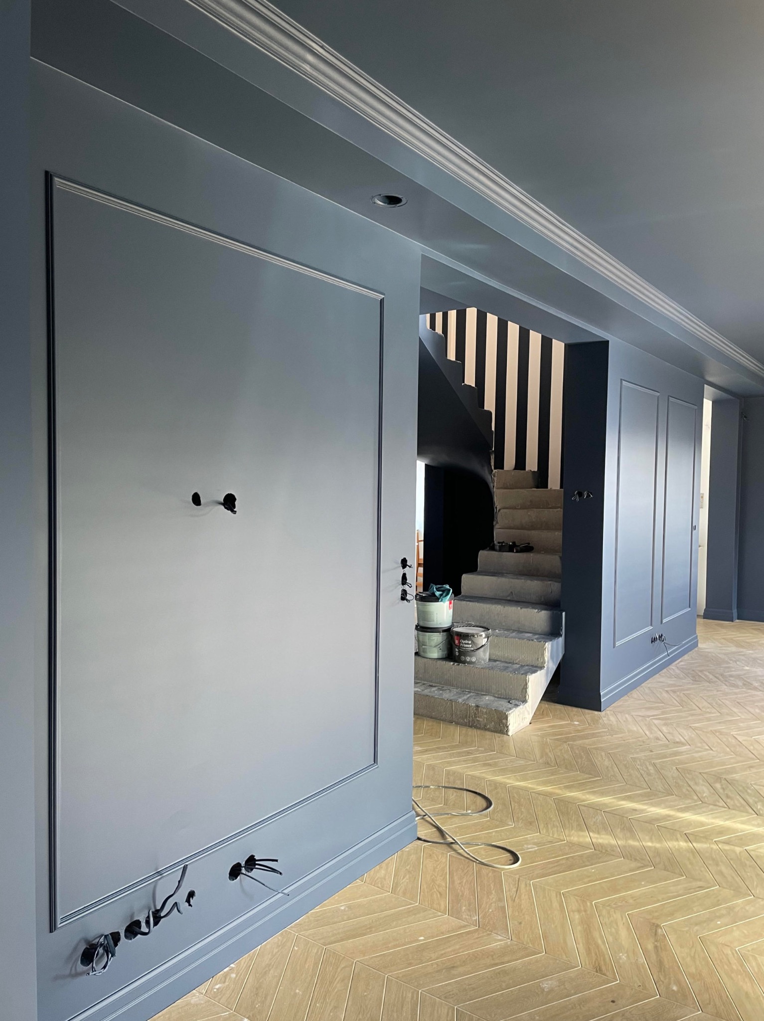 Świeżo pomalowane na szaro ściany z dekoracyjnymi listwami w eleganckim wnętrzu, widoczne puszki z farbą na surowych betonowych schodach z czarno-białymi pasami na ścianie klatki schodowej...