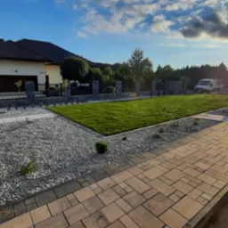 Nowo założony ogród z równo przystrzyżonym trawnikiem, ścieżką z kostki brukowej i dekoracyjnym żwirem, na tle domu jednorodzinnego i zachodzącego słońca.