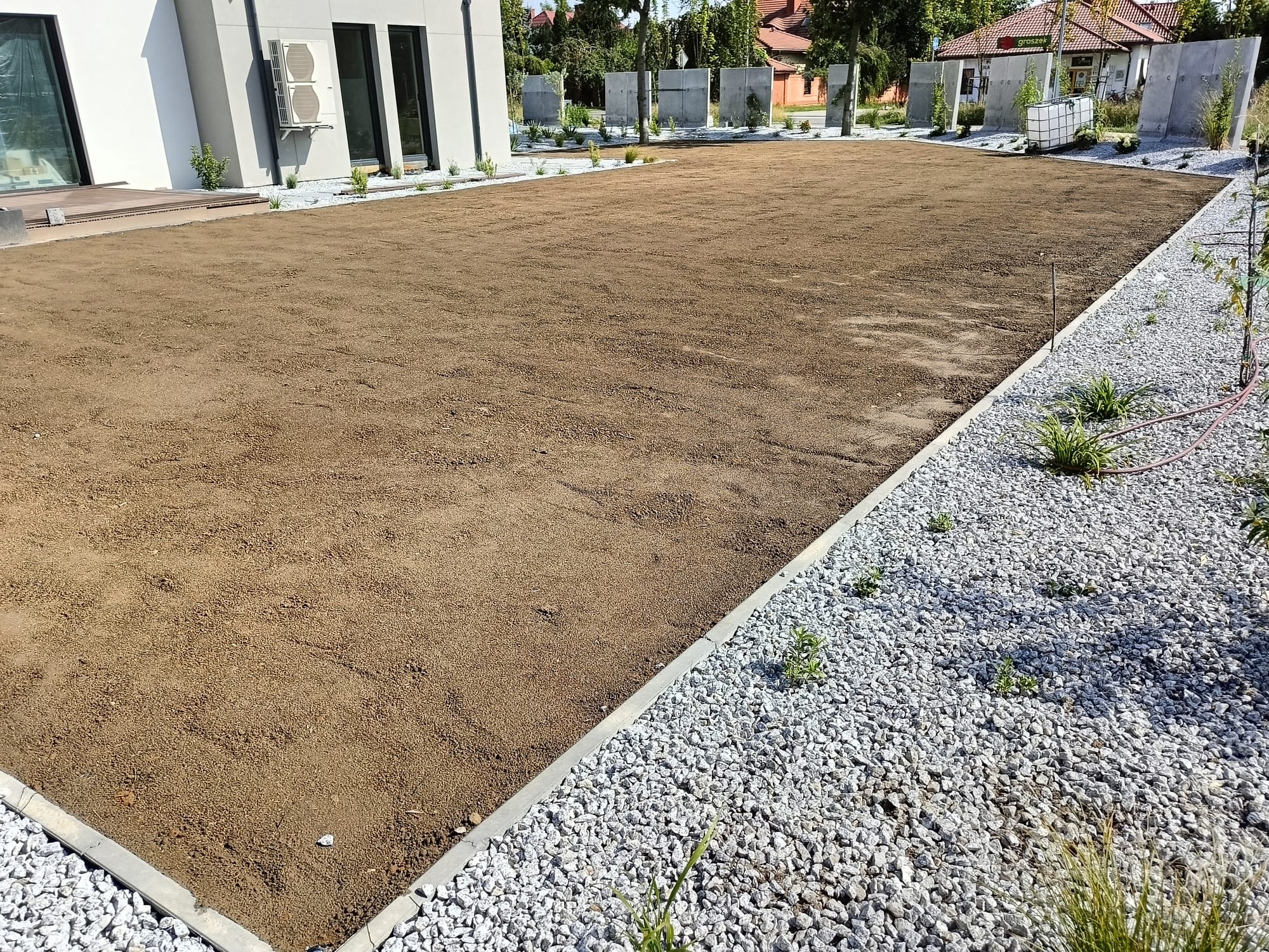 Przygotowany teren pod trawnik z nawiezioną ziemią, obrzeża wykończone kamieniami i betonowymi krawężnikami, widoczny nowoczesny dom z dużymi oknami i klimatyzatorem na elewacji.