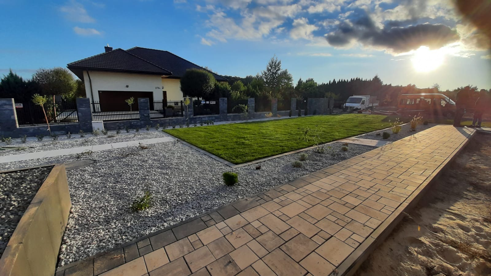 Nowo założony ogród z równo przystrzyżonym trawnikiem, ścieżką z kostki brukowej i dekoracyjnym żwirem, na tle domu jednorodzinnego i zachodzącego słońca.