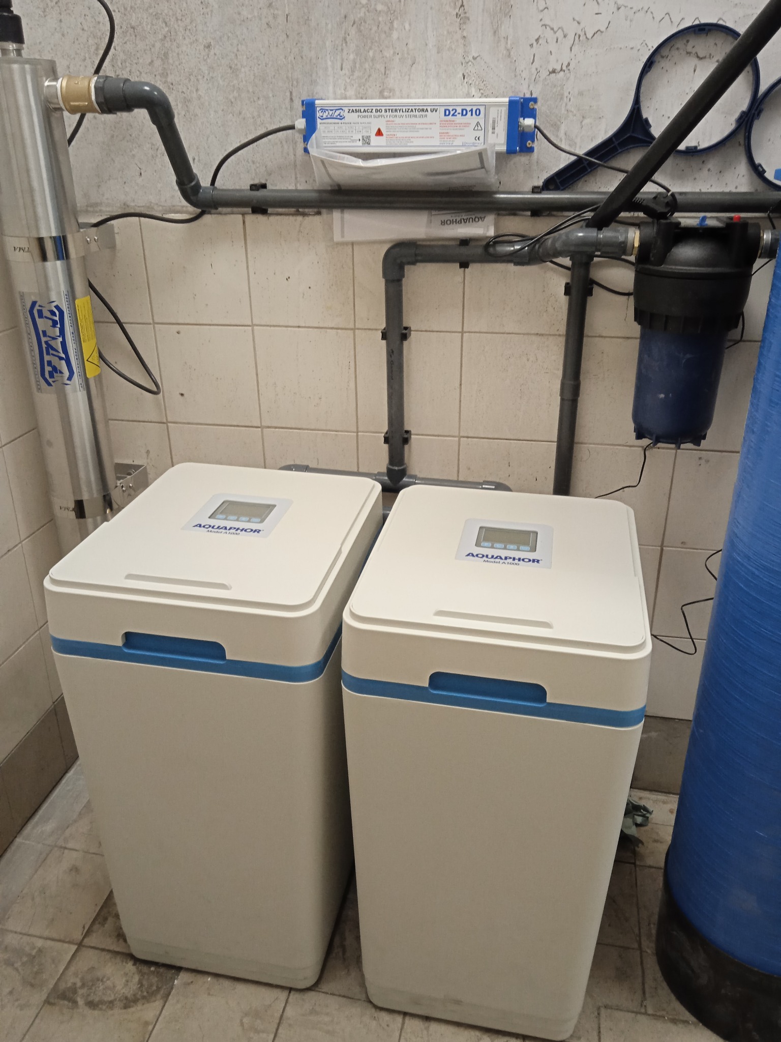Dwa białe filtry wody Aquaphor Model A1000 z niebieskimi akcentami, zainstalowane obok srebrnego sterylizatora UV i niebieskiego filtra wstępnego, z widocznymi rurami i kablami na tle kafelkowanej...