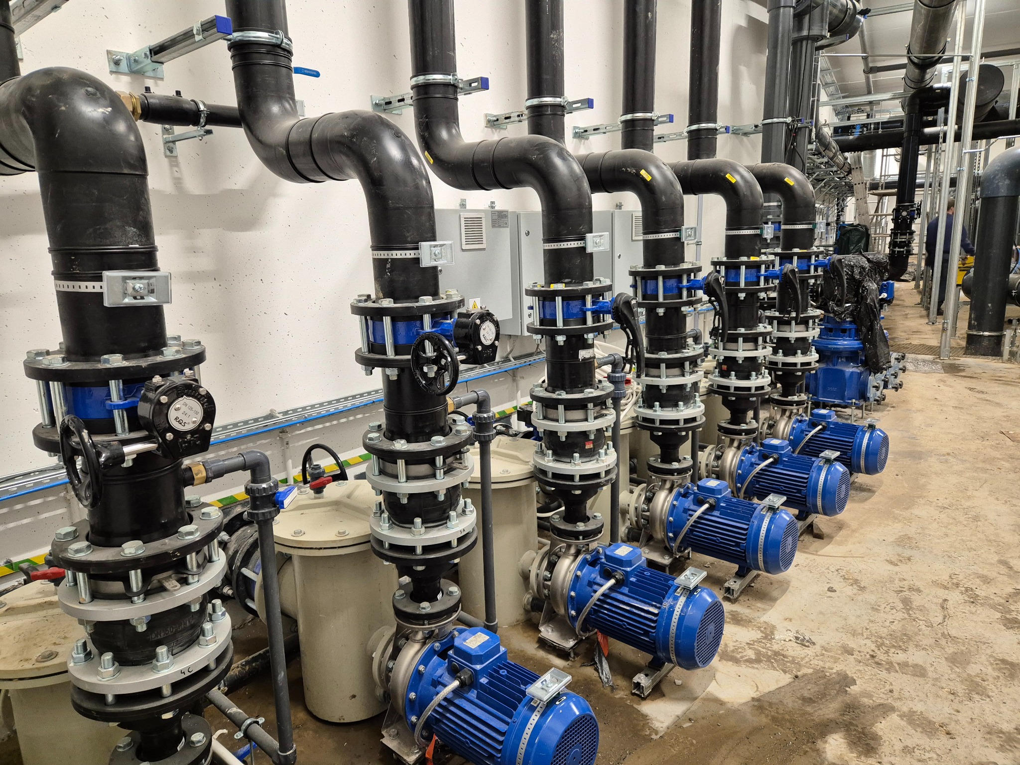 Rząd identycznych, niebieskich pomp przemysłowych z czarnymi rurami połączonych srebrnymi kołnierzami, instalacja w jasnym pomieszczeniu technicznym.