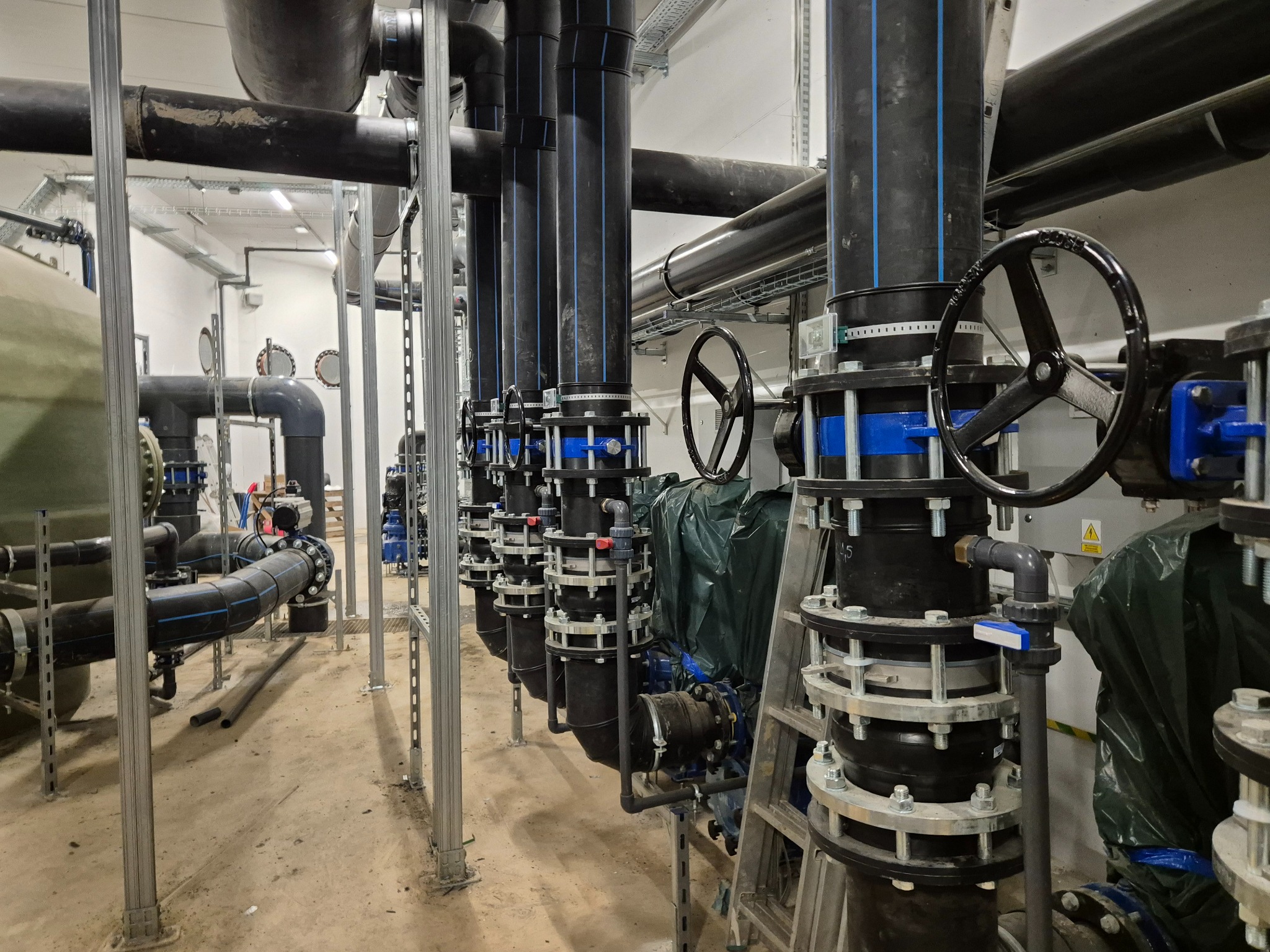System rur kanalizacyjnych z zaworami i elementami mocującymi w hali technologicznej, widok z dołu na czarne rury z niebieskimi paskami, metalowe podpory i zawory z kołami regulacyjnymi.