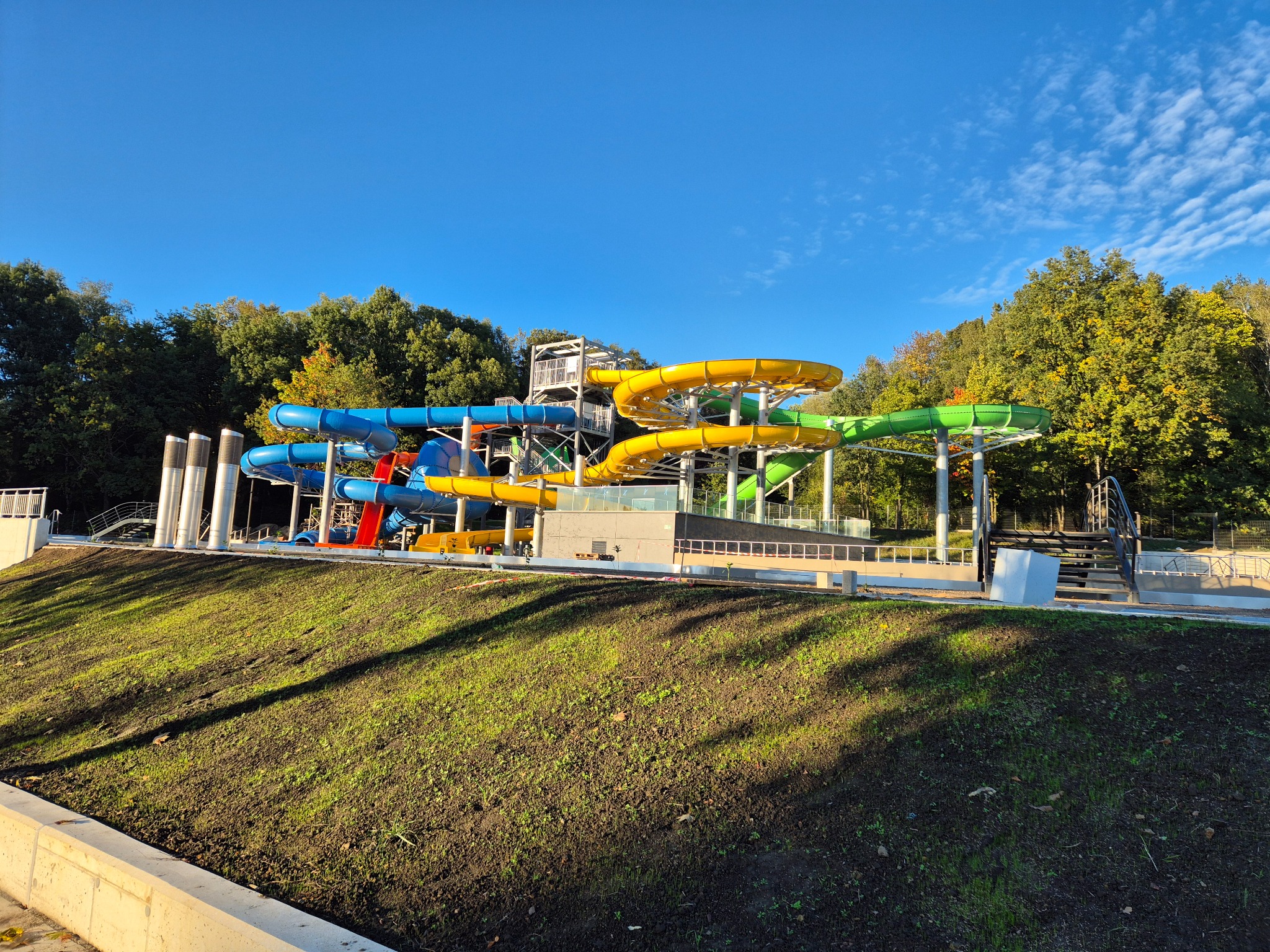 Kolorowy park wodny z licznymi zjeżdżalniami na tle zieleni drzew i błękitnego nieba, widoczny świeżo posadzony trawnik.