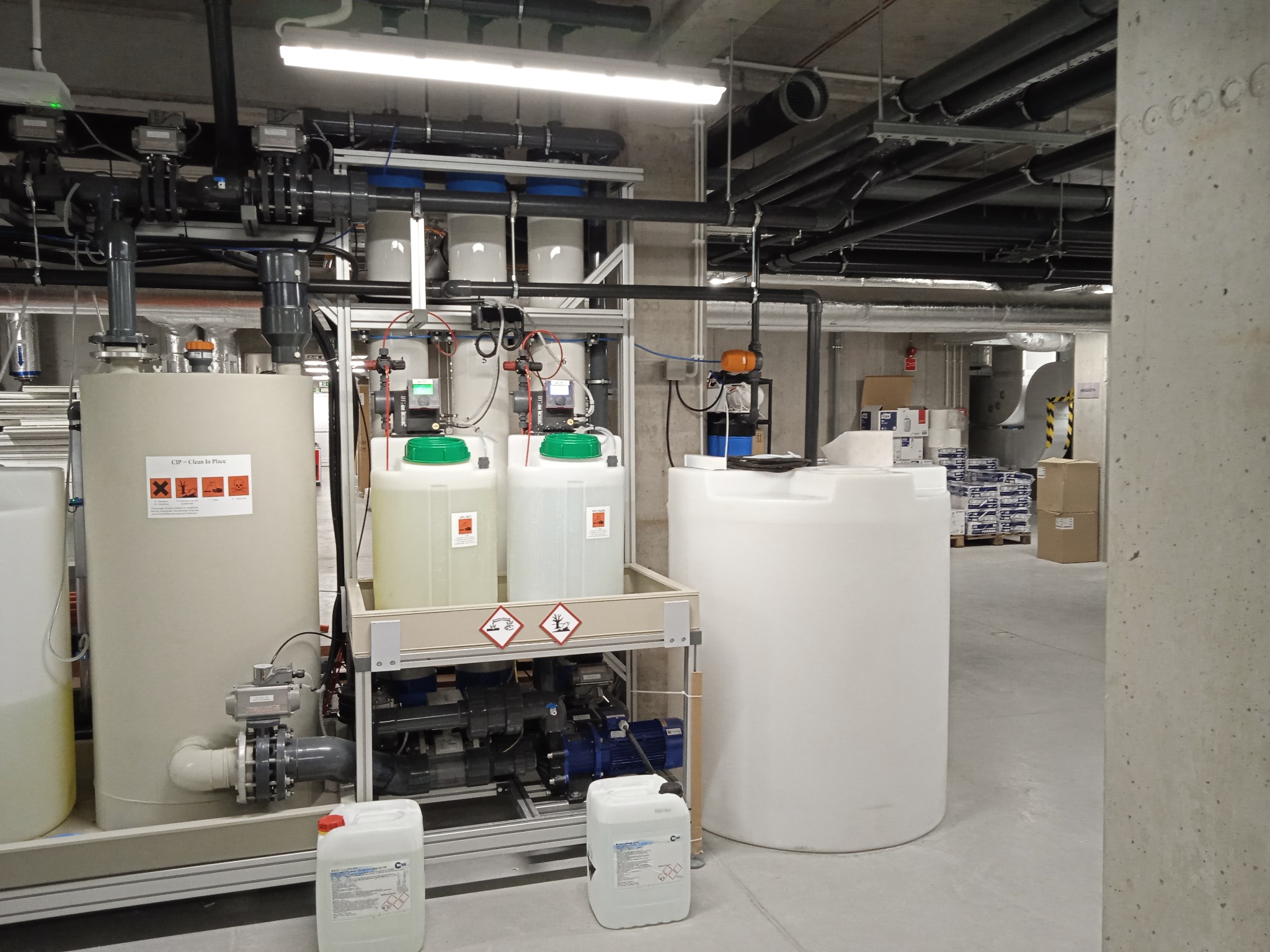Instalacja przemysłowa z białymi zbiornikami na ciecze, rurami i aparaturą kontrolno-pomiarową w surowym, betonowym pomieszczeniu. Widoczne znaki ostrzegawcze na zbiornikach. W tle kartony.