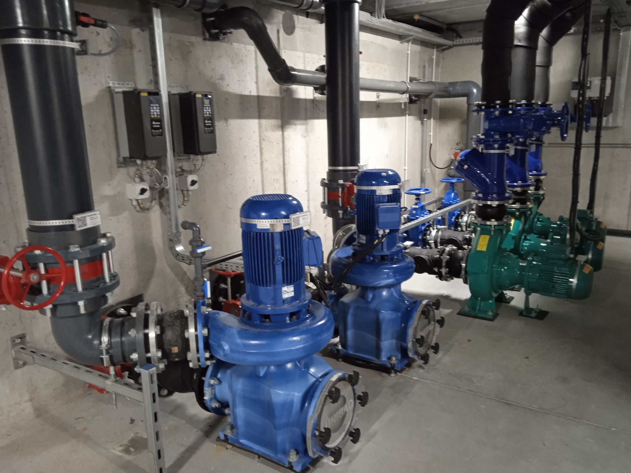 Instalacja pompowni z rurociągami, niebieskie i zielone pompy, szare rury, regulatory na ścianie w pomieszczeniu technicznym.