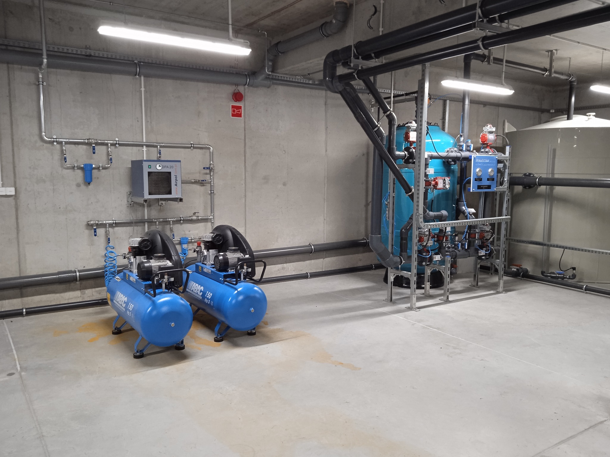 Kompletna instalacja przemysłowa z dwoma niebieskimi kompresorami ABAC 150, pionowym zbiornikiem i systemem rur na tle szarej ściany w hali.