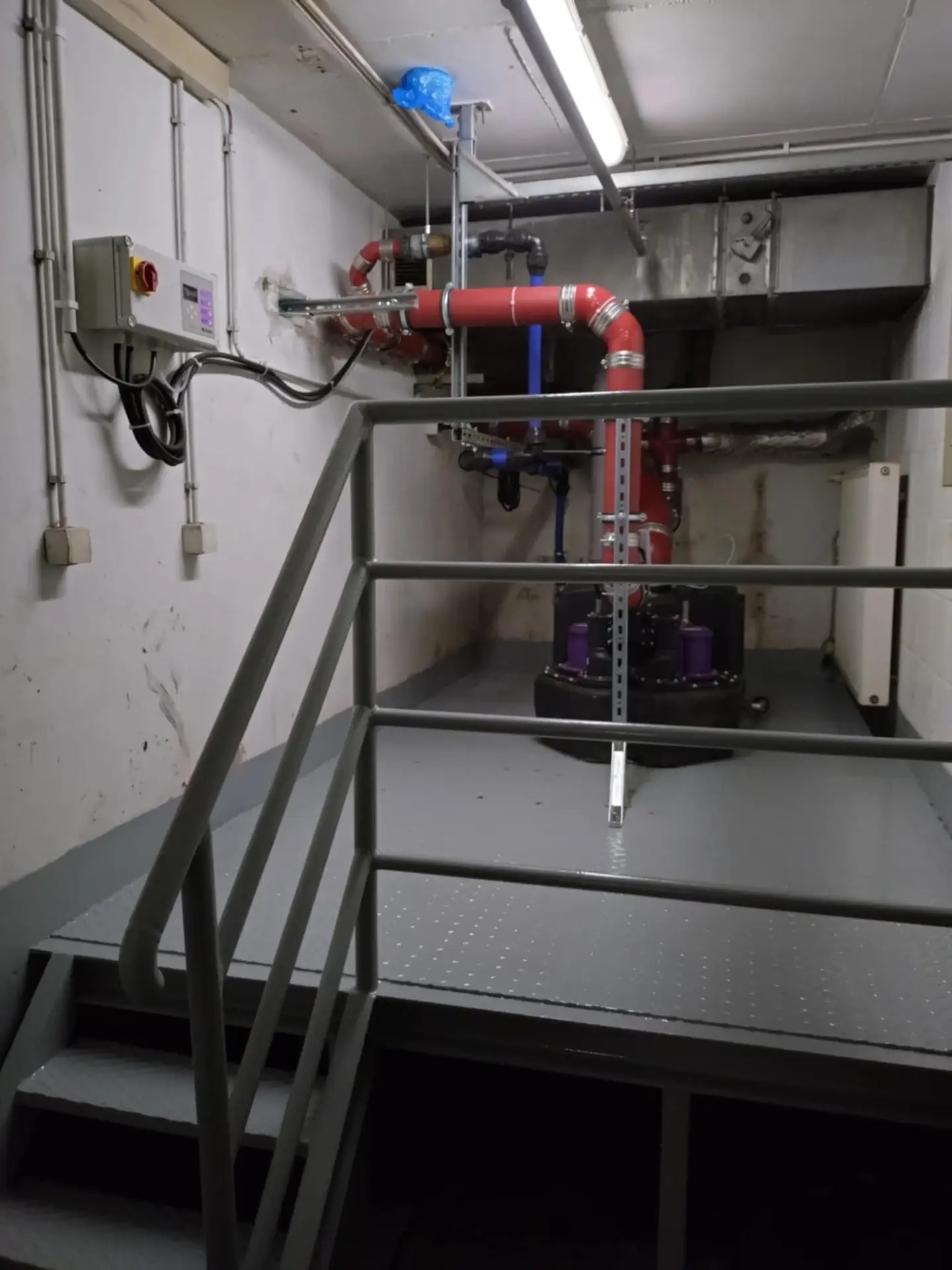 Wnętrze pomieszczenia technicznego z pompą zanurzeniową, rurociągami w kolorze czerwonym i niebieskim, szafą sterowniczą na ścianie i metalowymi schodami prowadzącymi na podest.