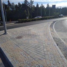 PPHU PIT-GRAN - Świeżo ułożona nawierzchnia z kostki brukowej w geometryczny wzór, obrzeża z kostki granitowej, widoczny właz kanalizacyjny, na drugim planie droga z samochodami i zieleń.