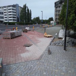 PPHU PIT-GRAN - Układanie czerwonej kostki brukowej i szarej kostki granitowej na placu budowy w mieście, widoczne palety z kostką, wózek paletowy i studzienka kanalizacyjna.