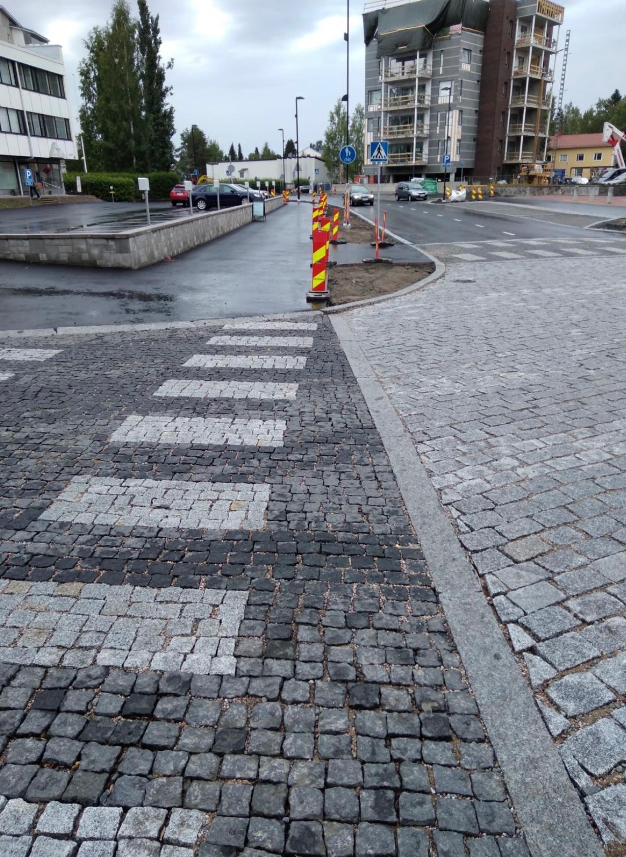 Ułożona kostka granitowa w formie przejścia dla pieszych z naprzemiennych szarych i czarnych kostek, obok nowa droga z kostki granitowej, w tle budynek w trakcie budowy i oznakowanie pionowe.