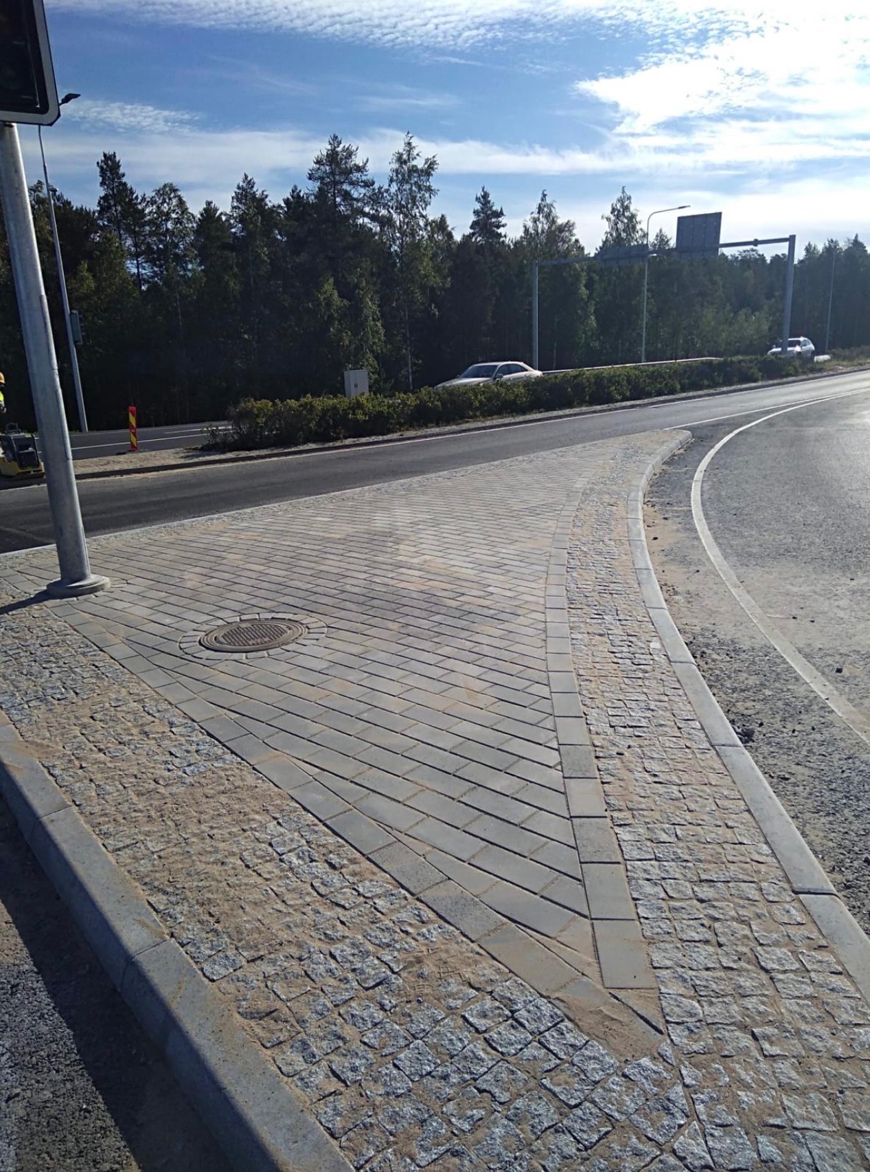 Świeżo ułożona nawierzchnia z kostki brukowej w geometryczny wzór, obrzeża z kostki granitowej, widoczny właz kanalizacyjny, na drugim planie droga z samochodami i zieleń.