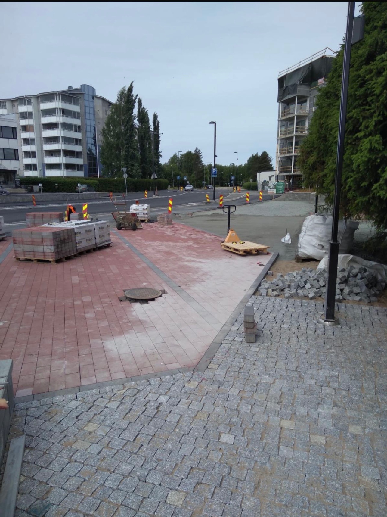 Układanie czerwonej kostki brukowej i szarej kostki granitowej na placu budowy w mieście, widoczne palety z kostką, wózek paletowy i studzienka kanalizacyjna.