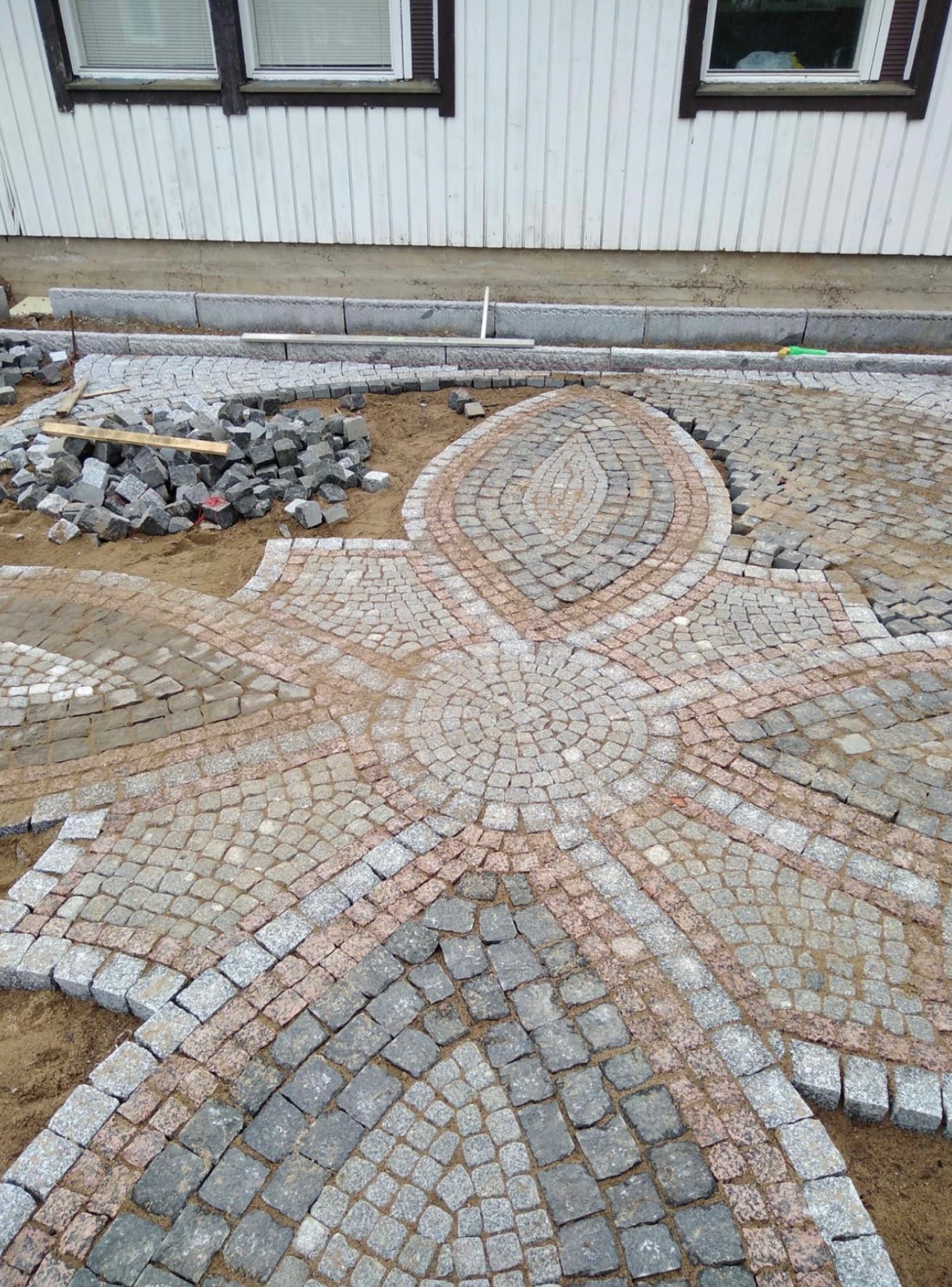 Fragment układanej nawierzchni z kostki granitowej w geometryczny wzór, widoczny stos kostki, obrzeża i fragment elewacji budynku w tle.