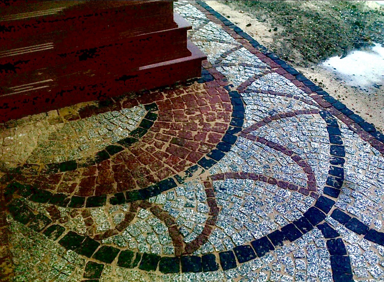 Wzór z kostki granitowej w odcieniach szarości, brązu i czerni, tworzący dekoracyjny motyw przed schodami wejściowymi.