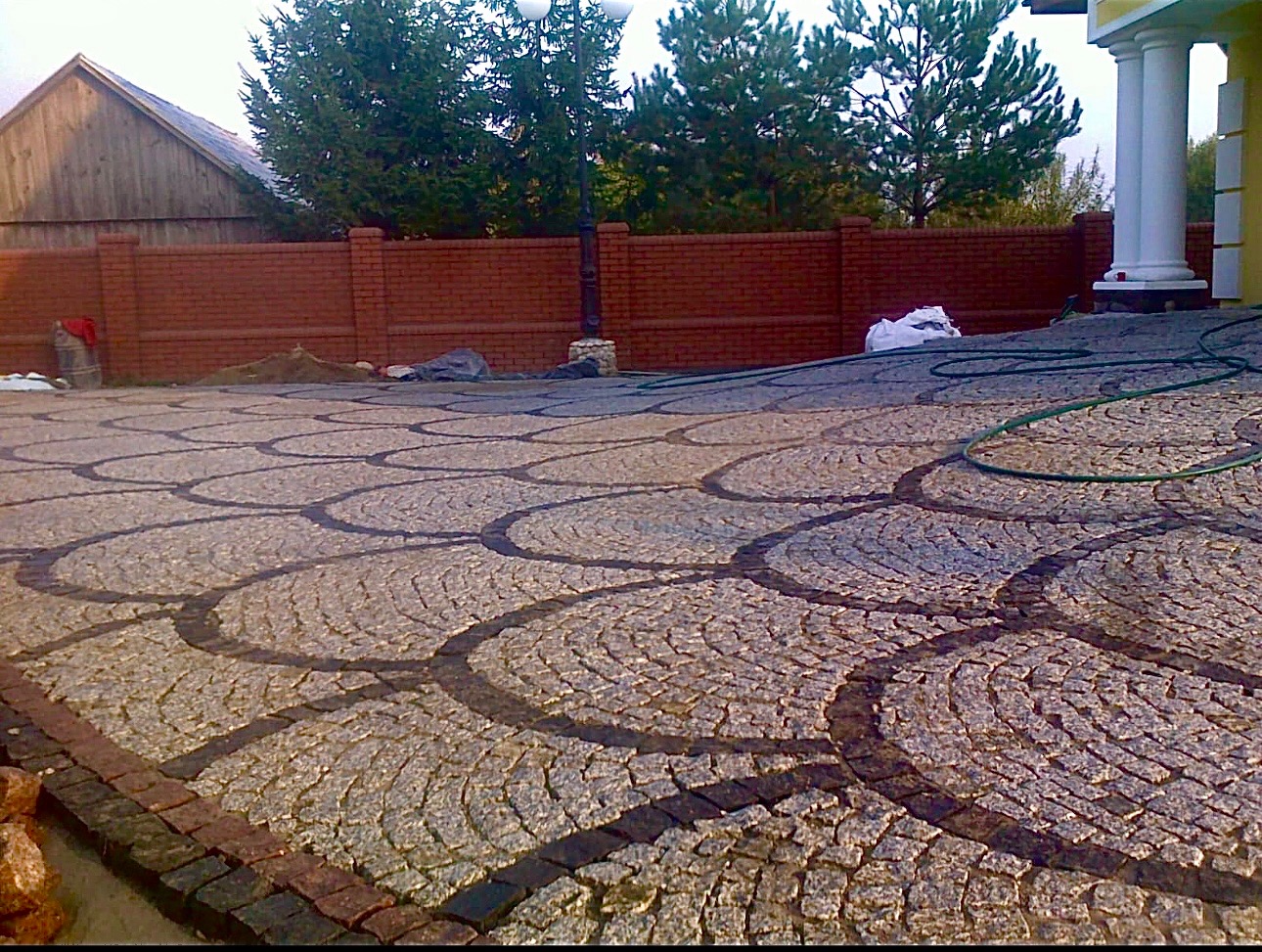 Ułożona kostka granitowa w wzór łusek ryby, obrzeże z ciemniejszej kostki, widoczny fragment domu z kolumnami i mur z cegły w tle.