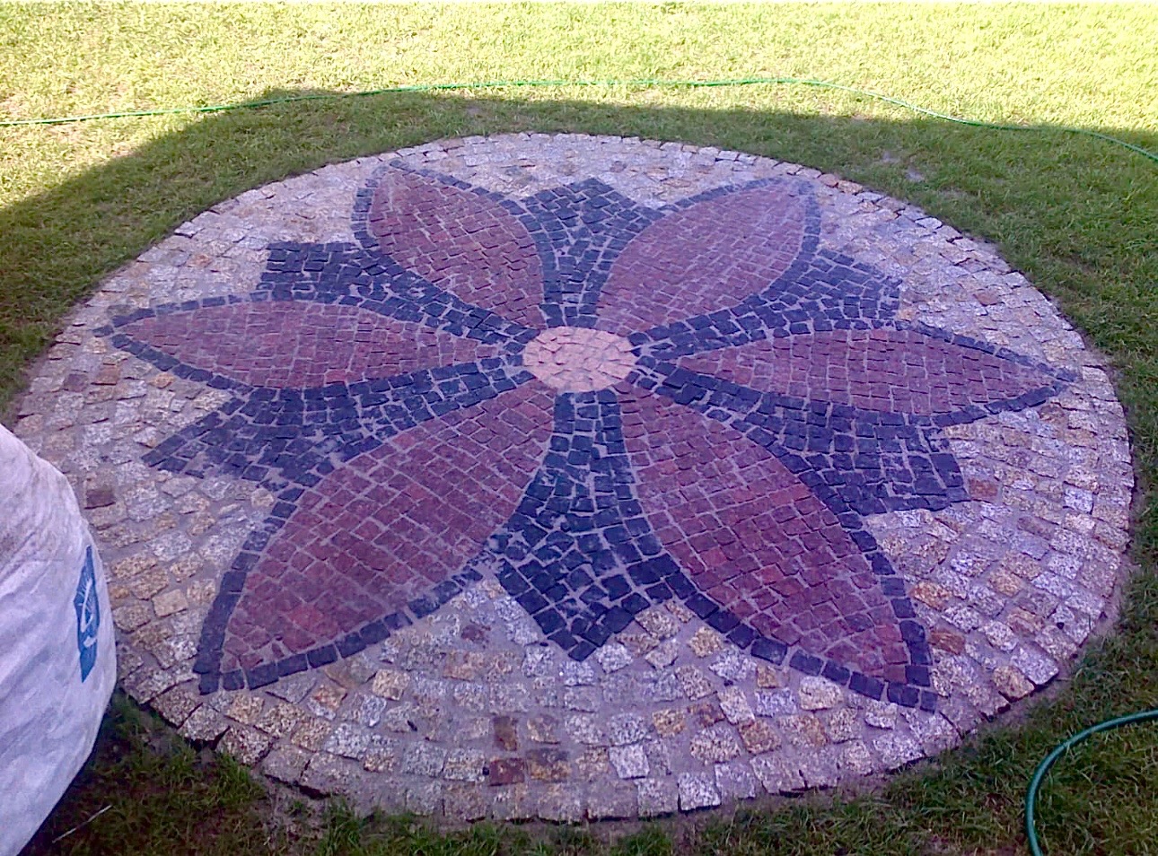 Okrągły wzór kwiatowy ułożony z kostki granitowej w Świdnicy, otoczony trawnikiem, z widocznym fragmentem worka z materiałem budowlanym.