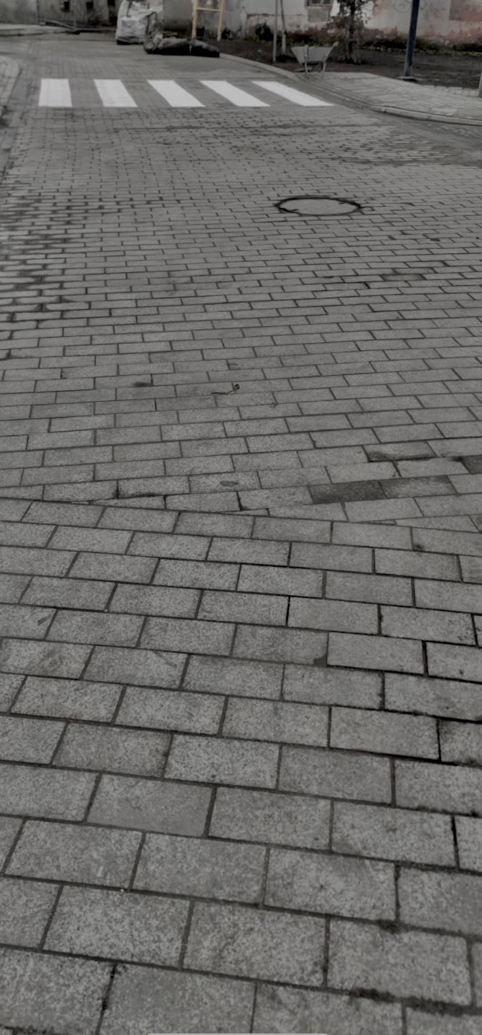 Szara kostka brukowa ułożona na ulicy z widocznym przejściem dla pieszych i studzienką kanalizacyjną, lekko wilgotna powierzchnia.