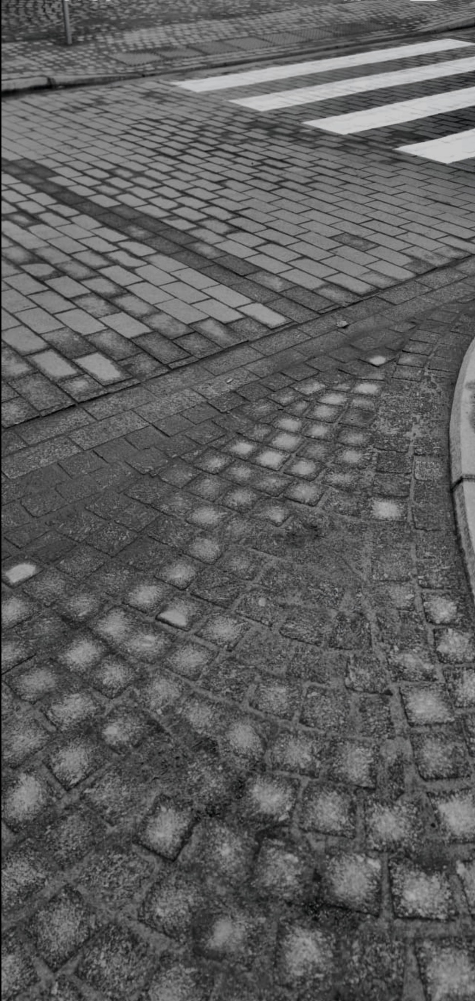 Ujęcie z góry na nawierzchnię z kostki brukowej, gdzie krzyżują się różne wzory ułożenia, a w tle widoczne przejście dla pieszych z białymi pasami.