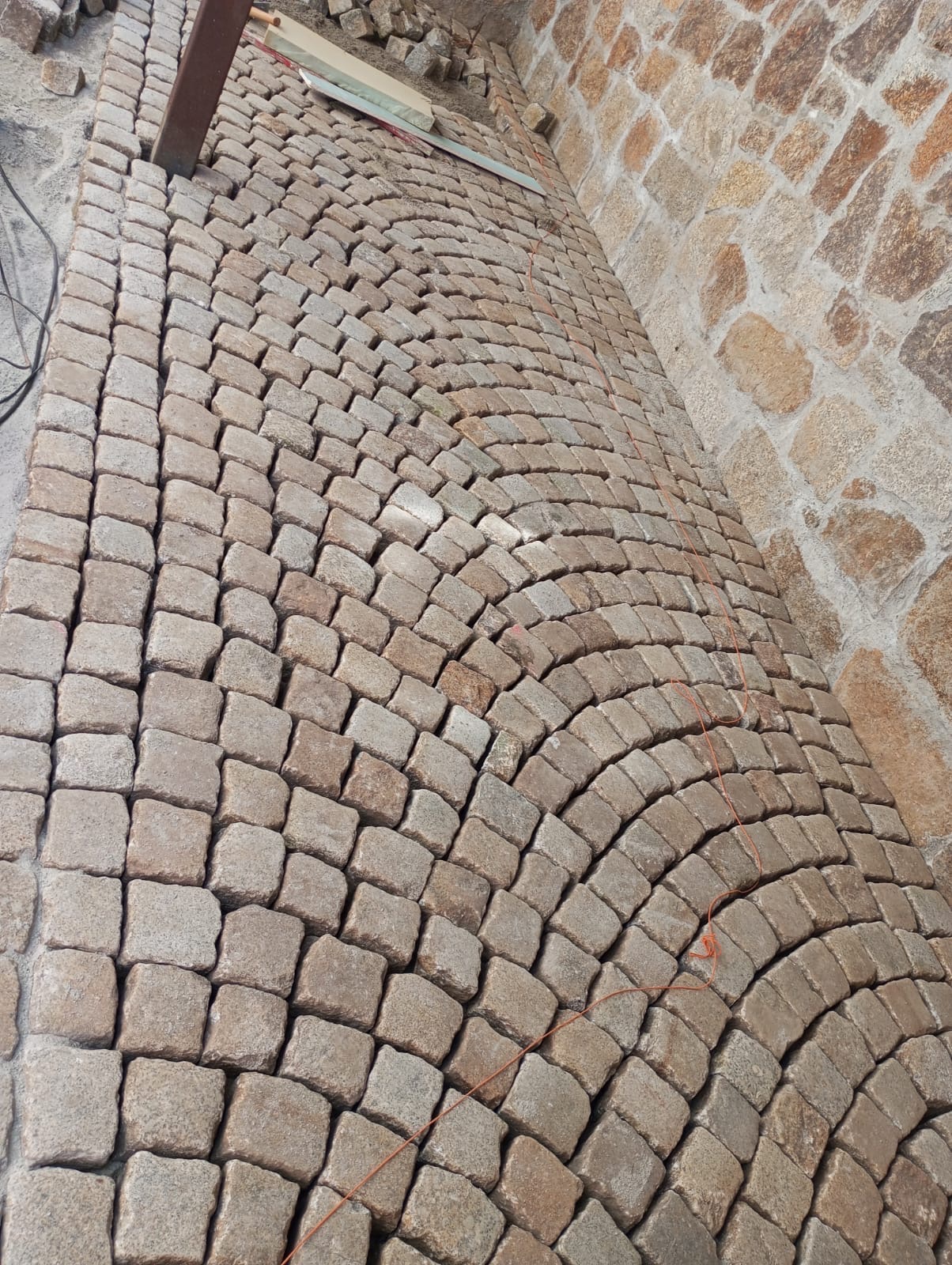 Ułożona kostka granitowa w łukowaty wzór przy kamiennej ścianie, z widocznymi narzędziami i resztkami materiału budowlanego.