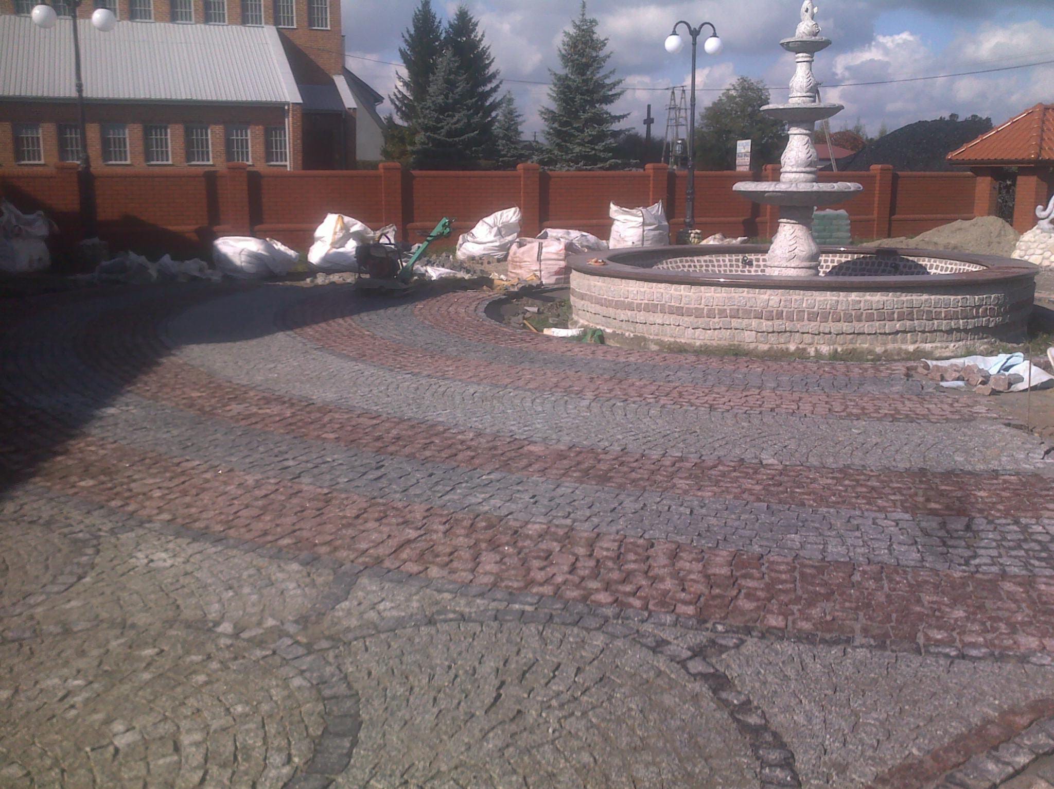 Plac z fontanną w trakcie wykańczania, z widocznym wzorem z kostki granitowej w odcieniach szarości i czerwieni, worki z materiałem budowlanym i narzędzia w tle.