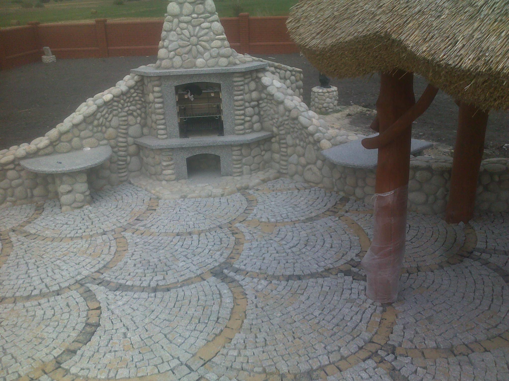 Ogród z paleniskiem i altaną, wykończony szarą kostką granitową ułożoną w półkoliste wzory z akcentami w kolorze piaskowym.