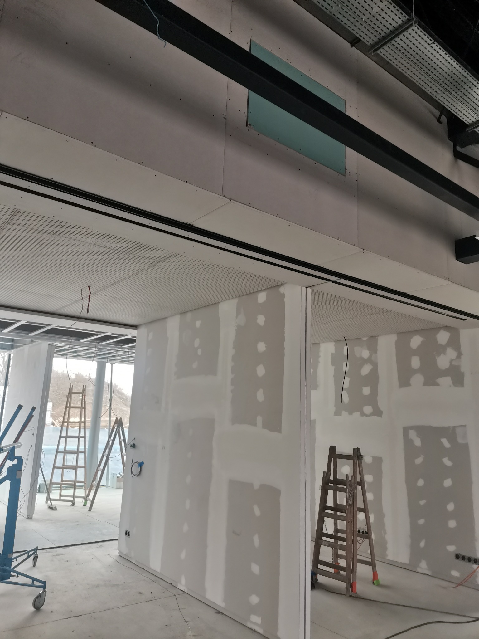 Wnętrze w trakcie prac wykończeniowych: ściany z płyt gipsowo-kartonowych z widocznymi miejscami po szpachlowaniu, drabiny, podnośnik do płyt, sufit podwieszany z perforacją, instalacje pod sufitem.