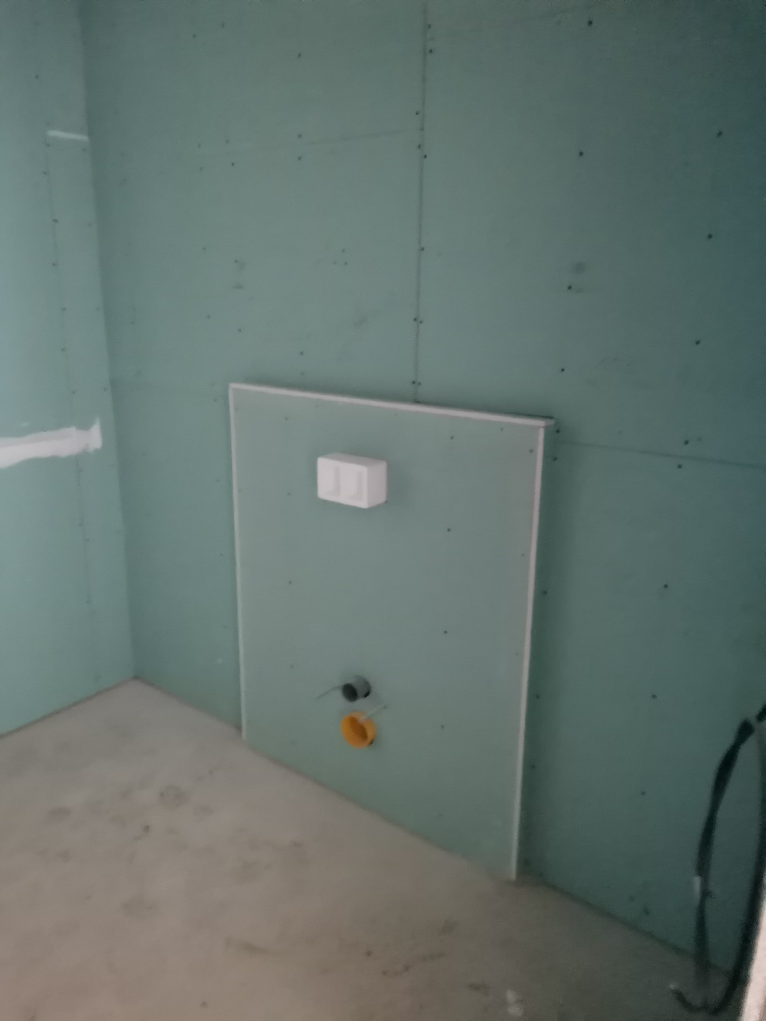 Niedokończona łazienka z zielonymi płytami gipsowo-kartonowymi, widoczne przyłącza do toalety podtynkowej i szpachlowane łączenia płyt.