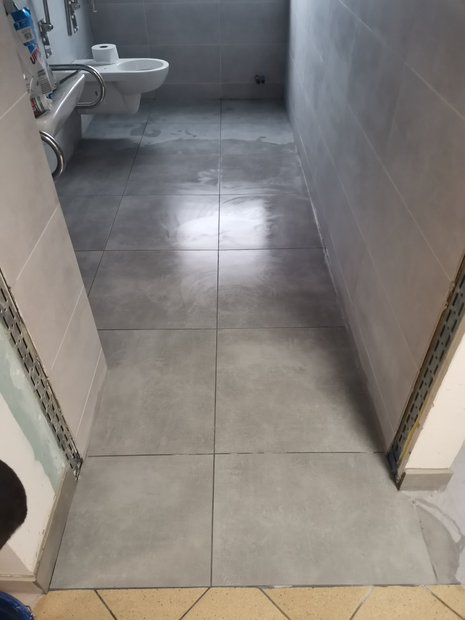 Szare płytki podłogowe i ścienne w małej łazience, widoczny fragment toalety i umywalki, listwa wykończeniowa przy drzwiach.