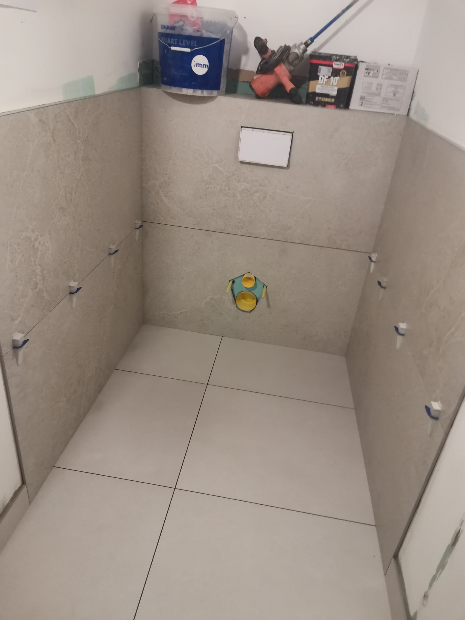 Wnętrze łazienki w trakcie kładzenia jasnych płytek ceramicznych na ścianach i podłodze, widoczne krzyżyki dystansowe, otwór instalacyjny, wiadro z zaprawą, mieszadło i opakowanie kleju na półce.