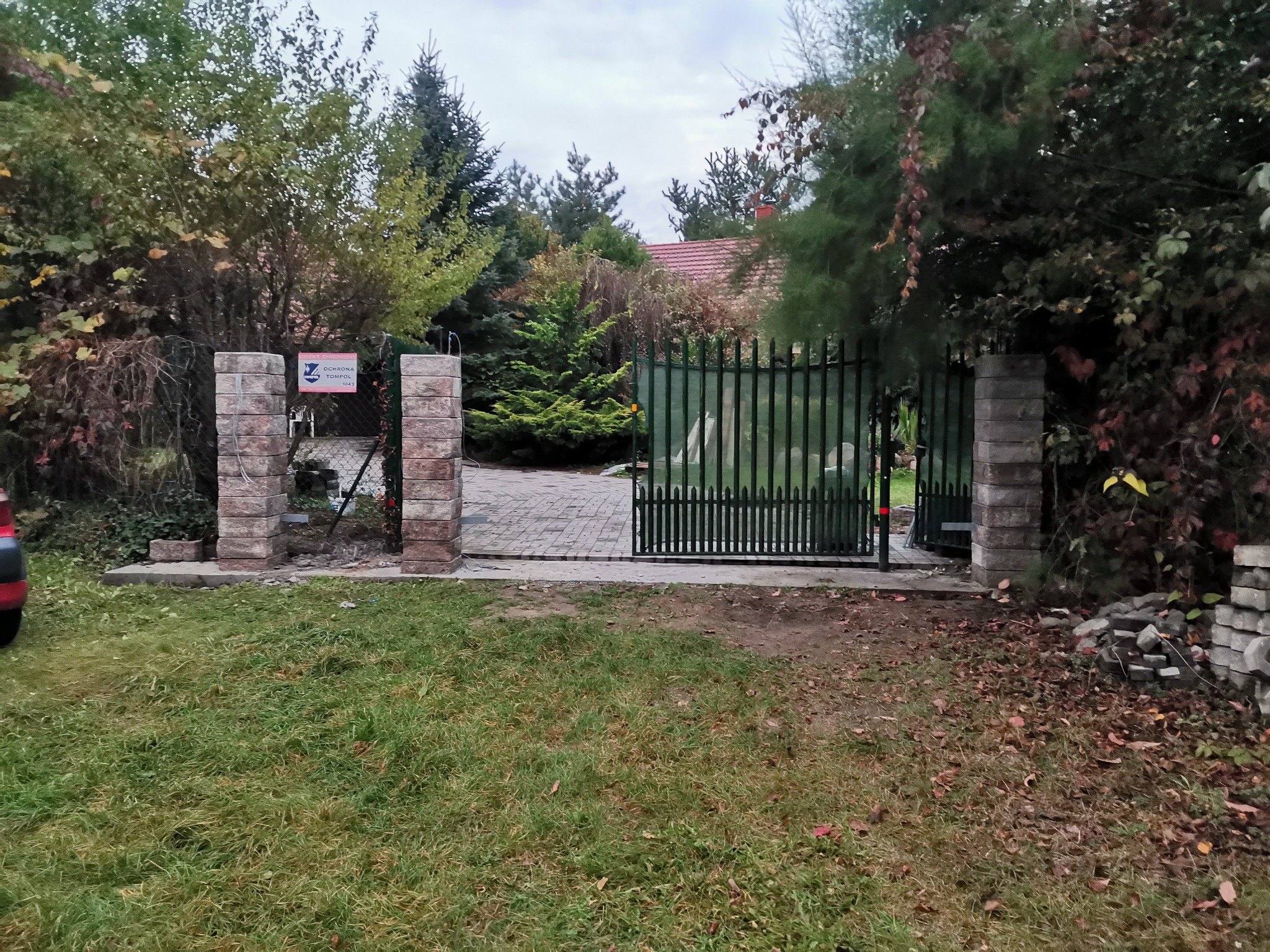 Fragment posesji z nowo wybudowaną bramą wjazdową z ceglanymi słupkami i metalowym ogrodzeniem, widok z zewnątrz.