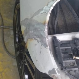 Yauheni Stayukhin - Fragment karoserii samochodu z widocznymi śladami spawania i szlifowania, przygotowany do dalszej obróbki lakierniczej, widoczne elementy zawieszenia i opona.