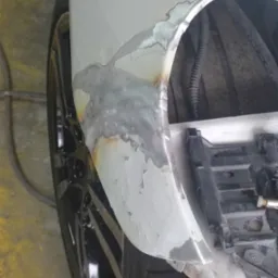 Fragment karoserii samochodu z widocznymi śladami spawania i szlifowania, przygotowany do dalszej obróbki lakierniczej, widoczne elementy zawieszenia i opona.