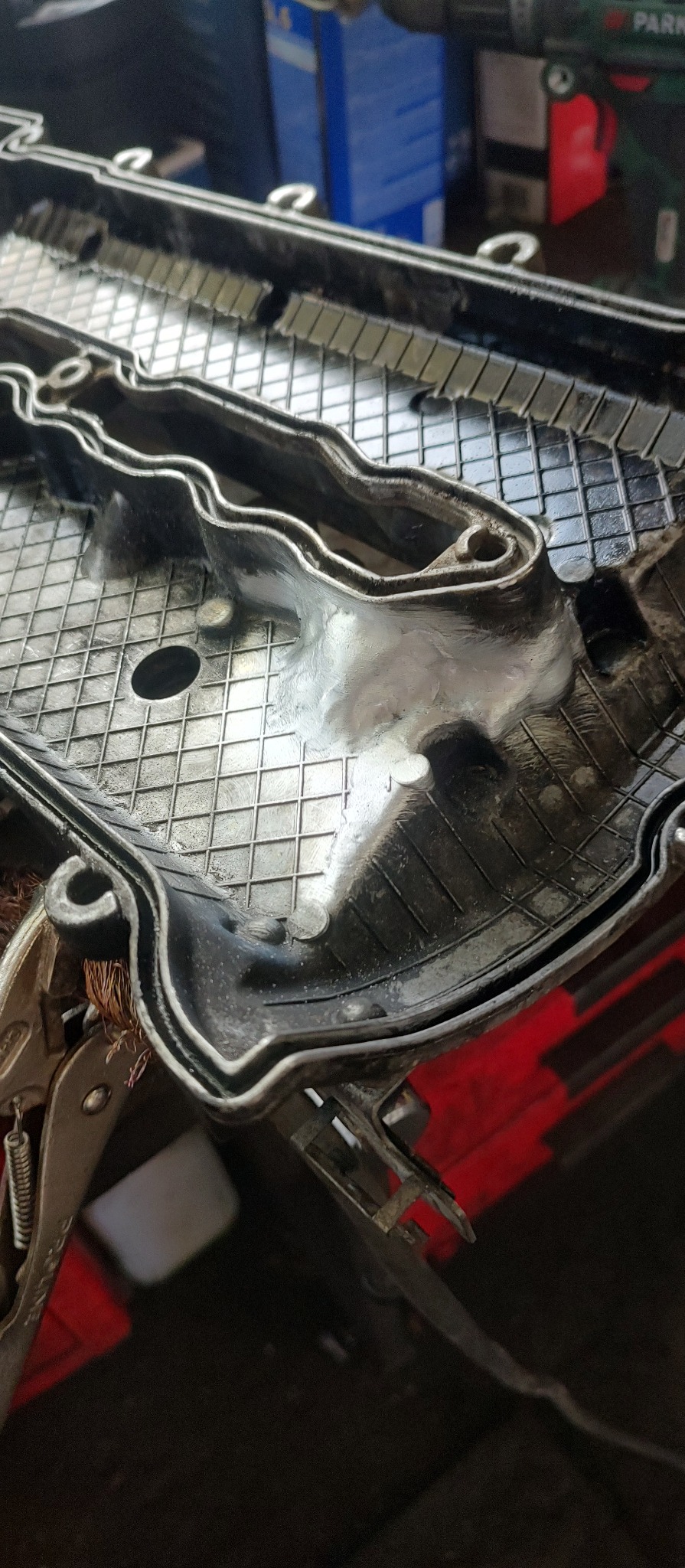 Naprawiona spawem pokrywa zaworów silnika samochodowego, widoczne ślady spawania aluminium i zacisk śrubowy.