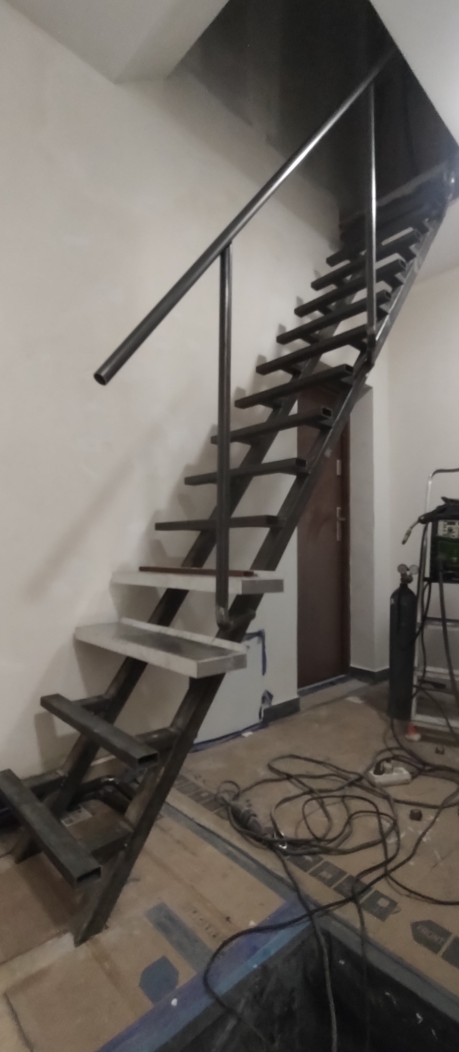 Metalowe schody z rur kwadratowych i poręczą, częściowo wykończone, widoczne narzędzia spawalnicze w tle.