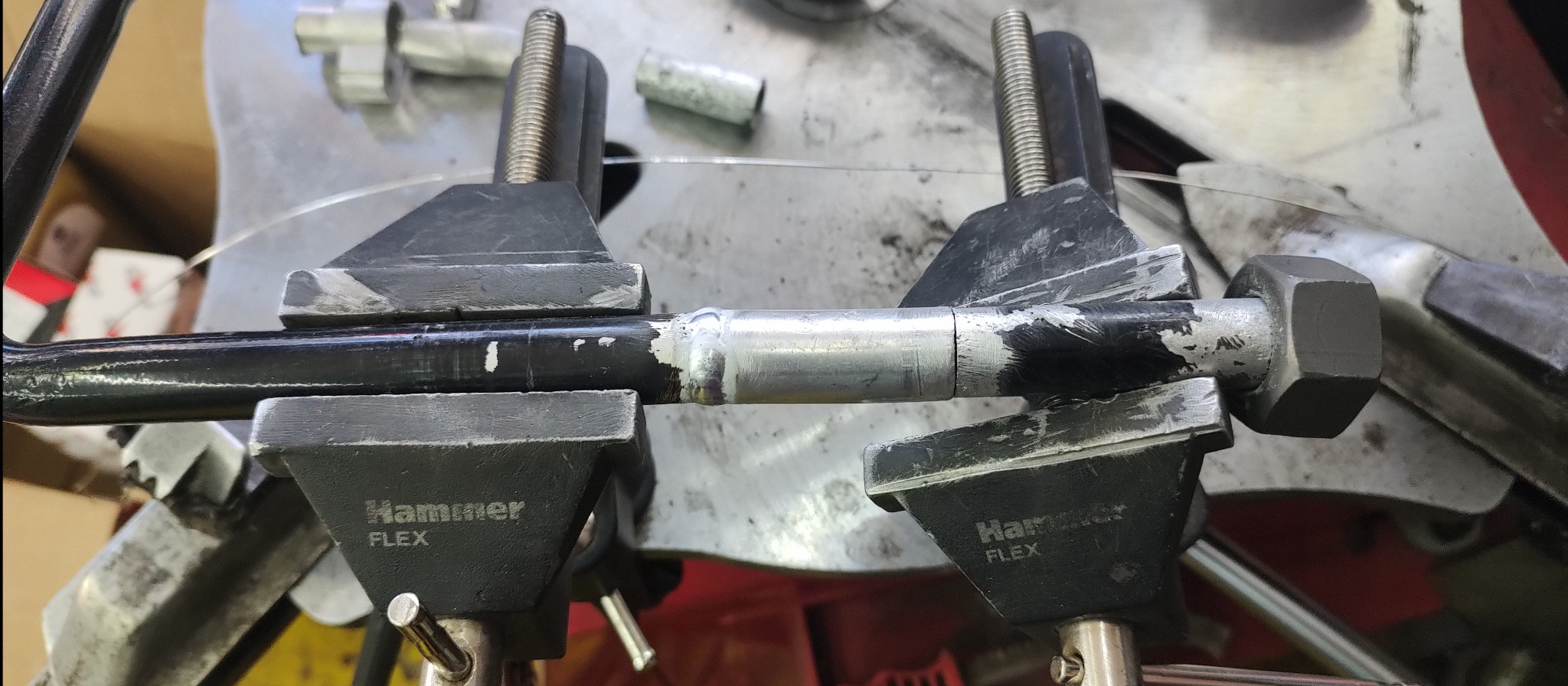 Metalowy pręt z widocznym spawem, zamocowany w dwóch imadłach marki Hammer Flex, ukazujący proces obróbki.