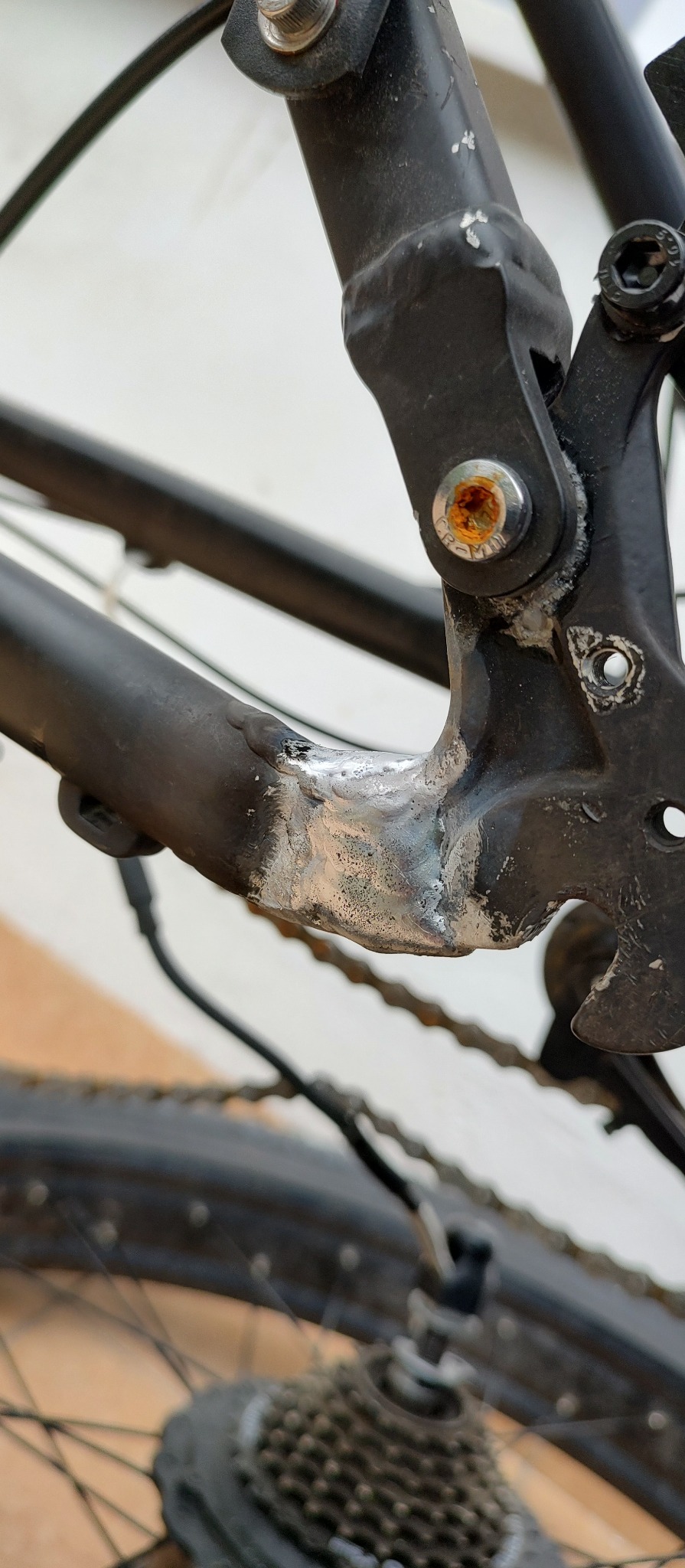 Szczegółowe ujęcie ramy roweru z widocznym spawem w miejscu łączenia elementów, śruby z rdzą i łańcuchem, wskazujące na naprawę lub wzmocnienie konstrukcji.