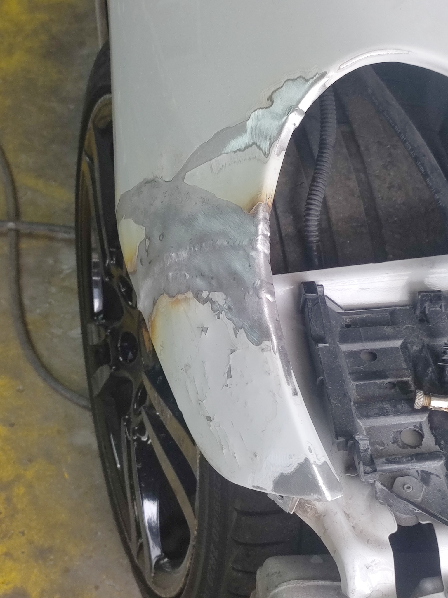 Fragment karoserii samochodu z widocznymi śladami spawania i szlifowania, przygotowany do dalszej obróbki lakierniczej, widoczne elementy zawieszenia i opona.